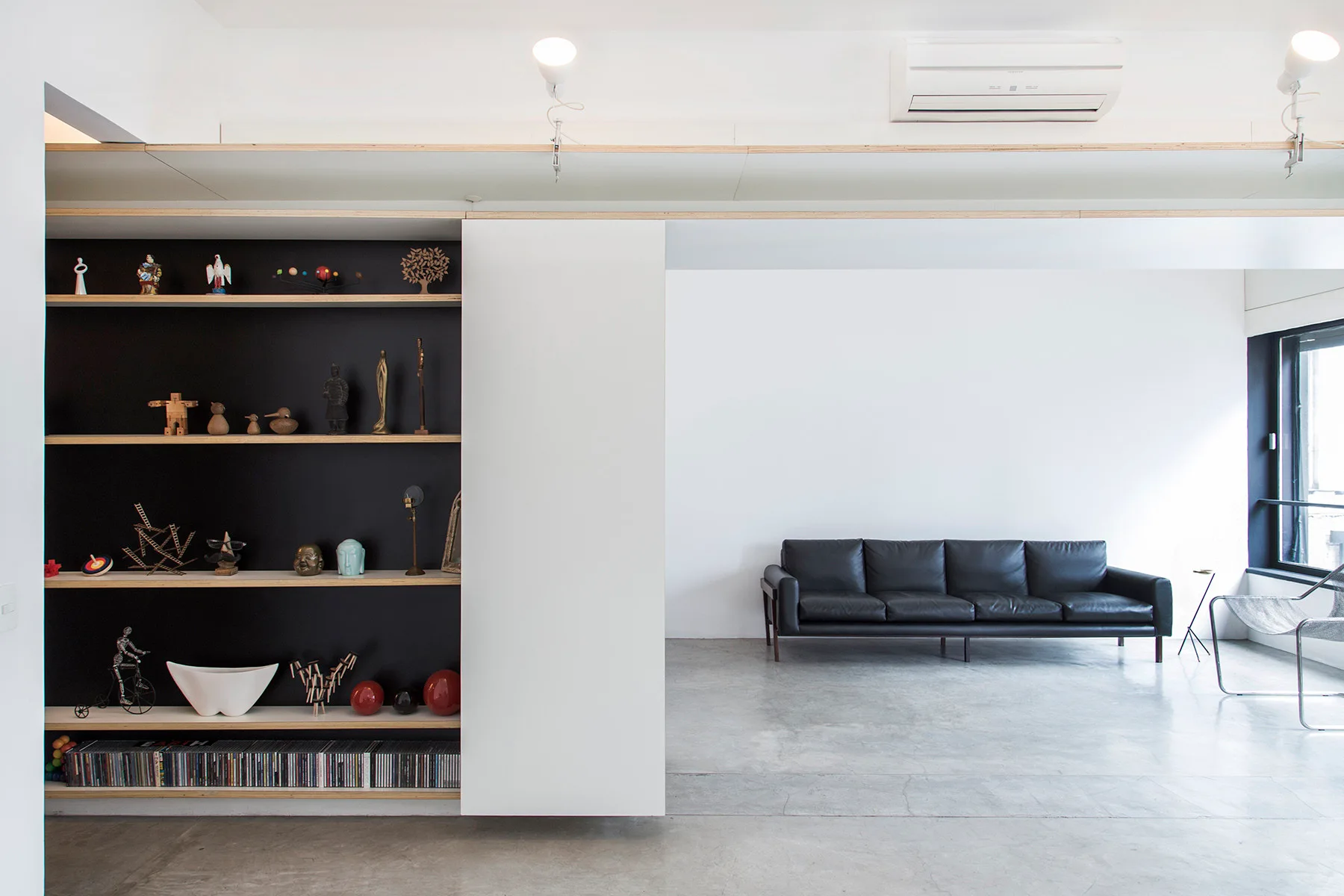 A minimalist living room features a black leather sofa, a smooth grey concrete floor, and a large black built-in shelving unit with a sliding white door.