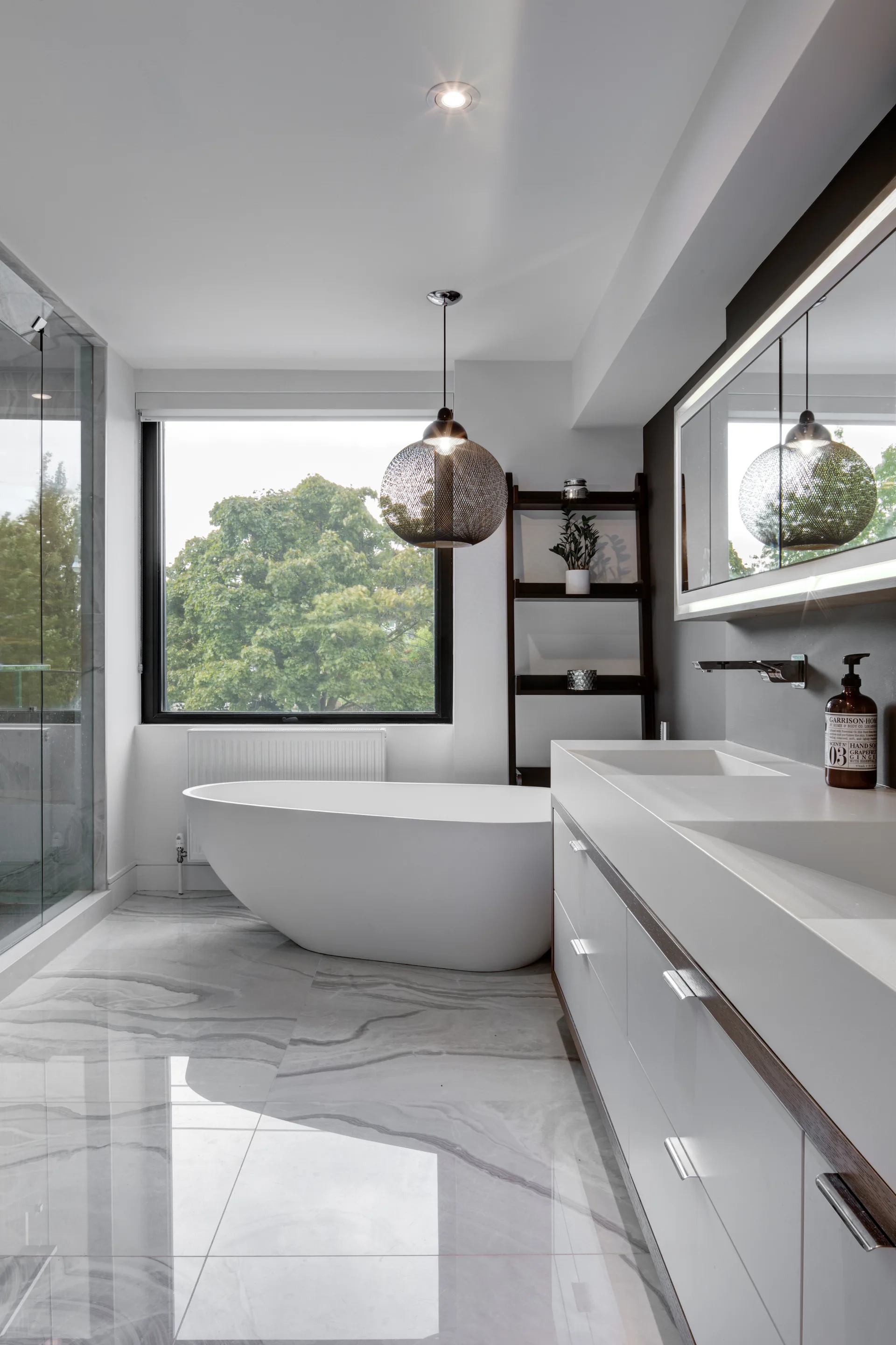 A modern bathroom features a white freestanding bathtub under a large window, a double vanity, and glossy veined tile flooring.