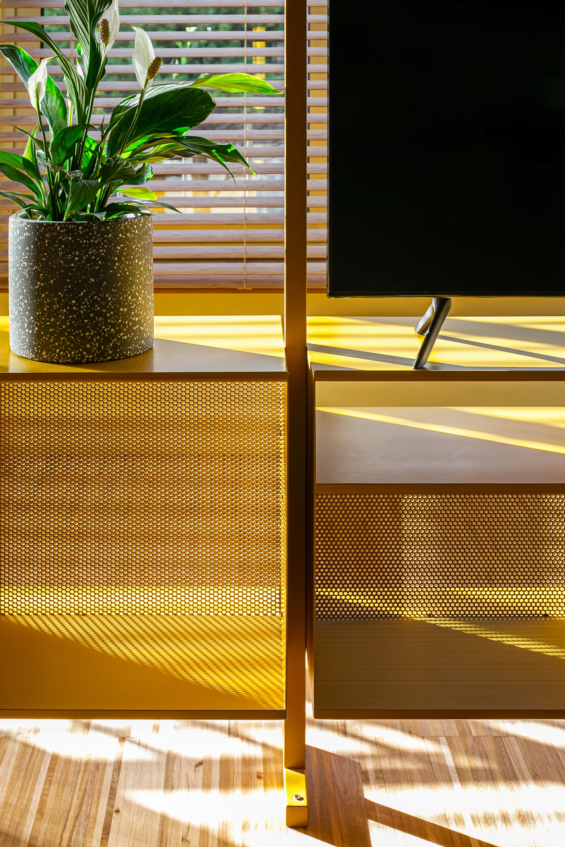 A yellow metal shelving unit features a speckled ceramic planter with a green plant and a television screen against a window with wooden blinds.
