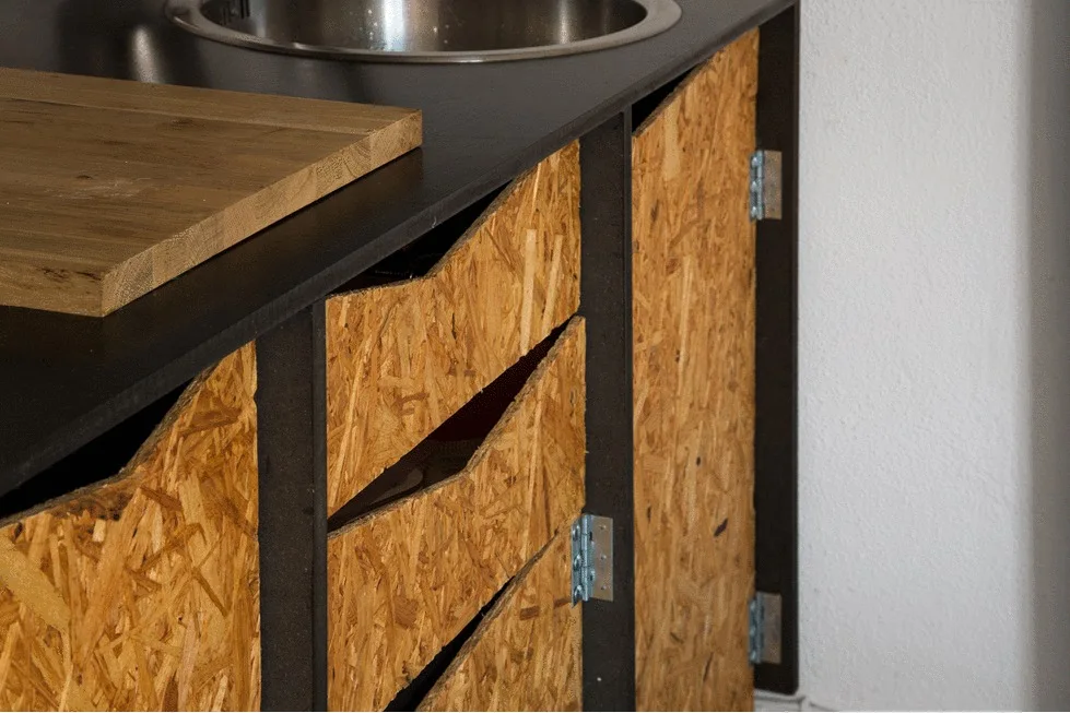 Close-up of kitchen cabinets featuring textured oriented strand board fronts, exposed metal hinges, and a black countertop with an integrated sink.