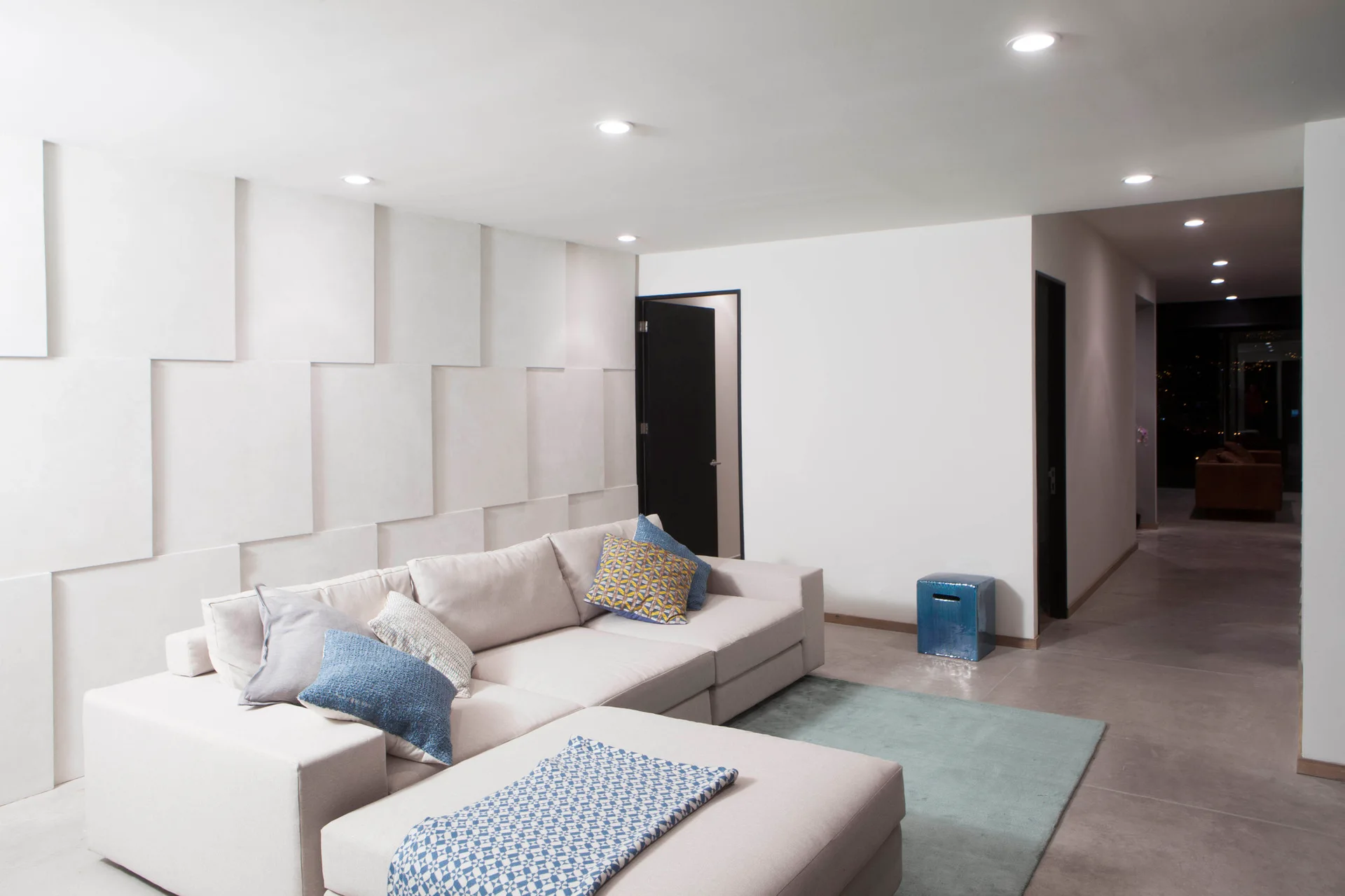 A beige sectional sofa rests against a white geometric paneled wall in a living room with a light blue rug and a glossy blue ceramic stool.