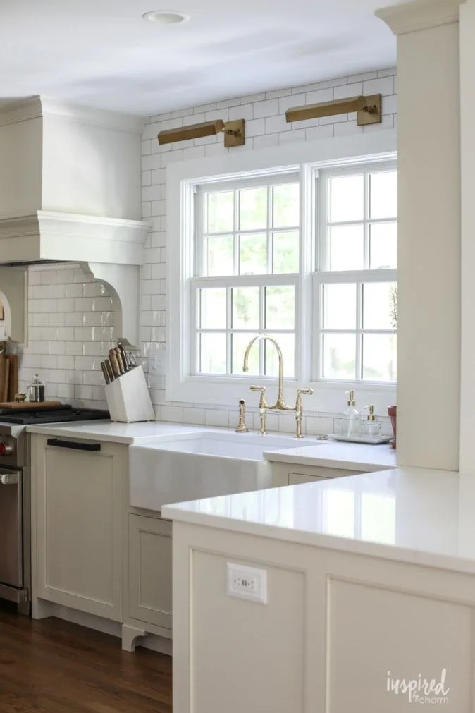 A white kitchen features a farmhouse sink under a double window, highlighted by a brass bridge faucet and two gold-toned picture lights on a subway tile wall.