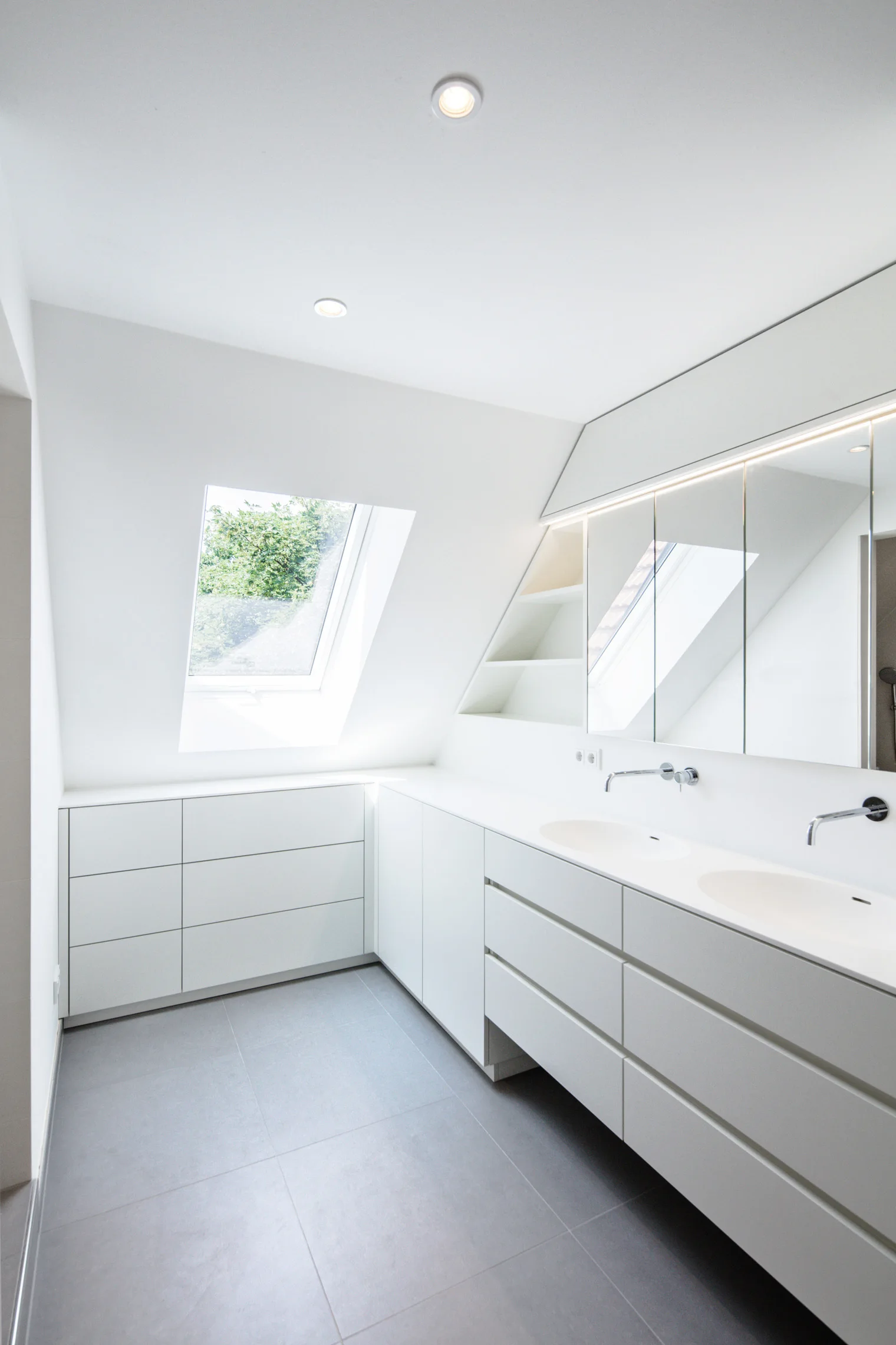 A minimalist white attic bathroom featuring a double vanity, wall-mounted faucets, built-in storage cabinets, and a slanted ceiling with a skylight.