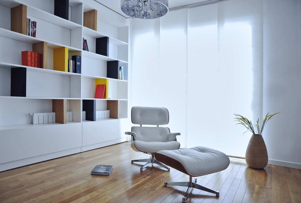 A minimalist room features a white leather lounge chair and ottoman on hardwood floors next to a large white built-in bookshelf and diffused panel blinds.
