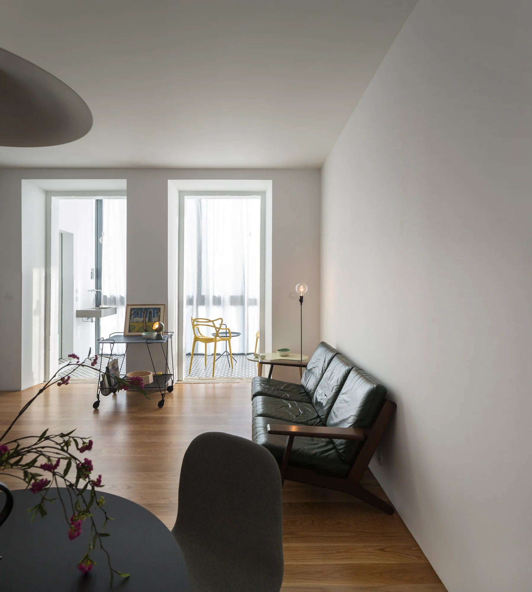 A dark green leather sofa rests on wood flooring in a minimalist living room next to large windows that lead to a tiled balcony.