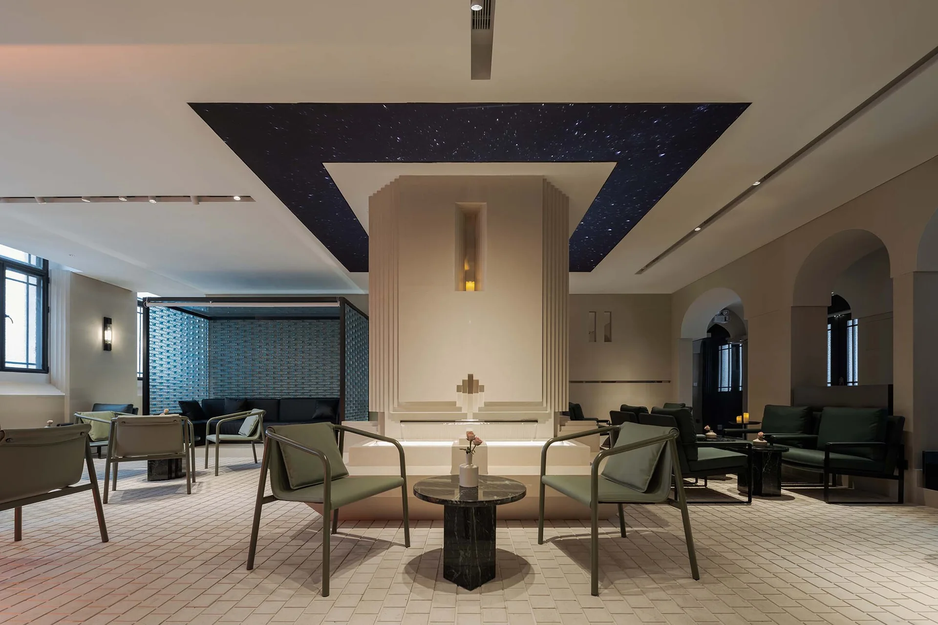 A contemporary lounge space features sage green armchairs arranged around a central beige fireplace column under a dark ceiling panel with tiny light points.