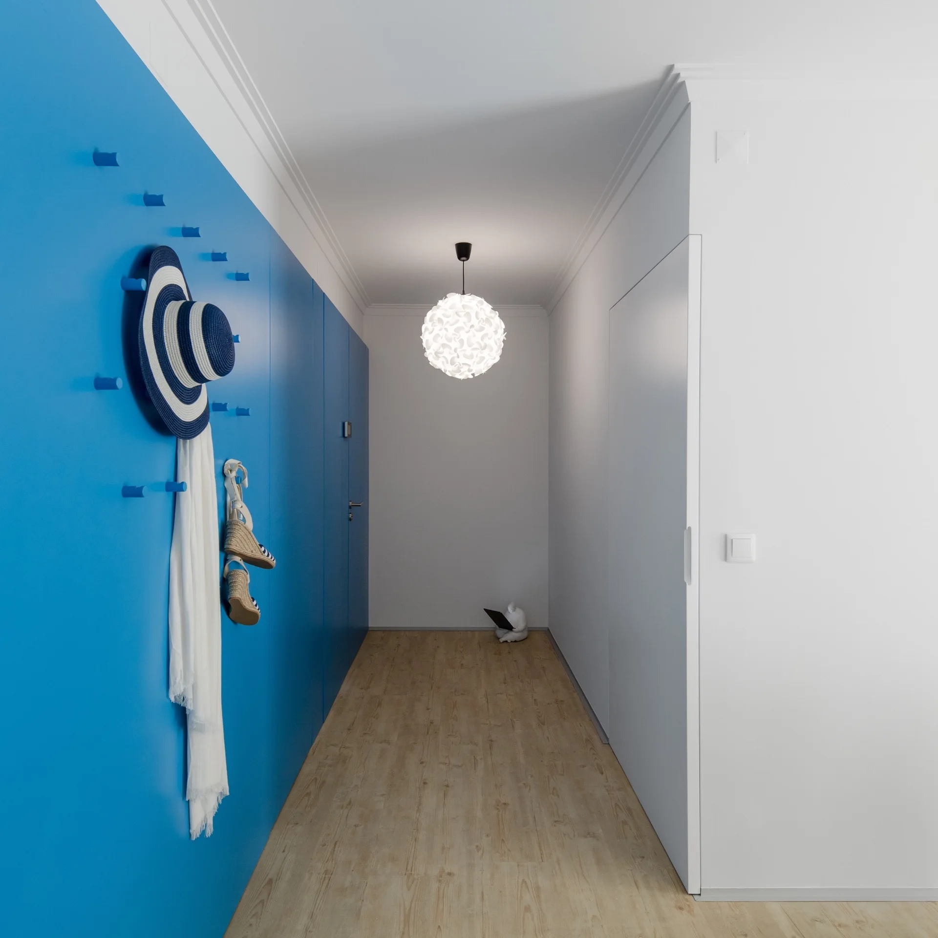 A minimalist white hallway features a vibrant blue accent wall with matching coat pegs, light wood flooring, and a round pendant light.