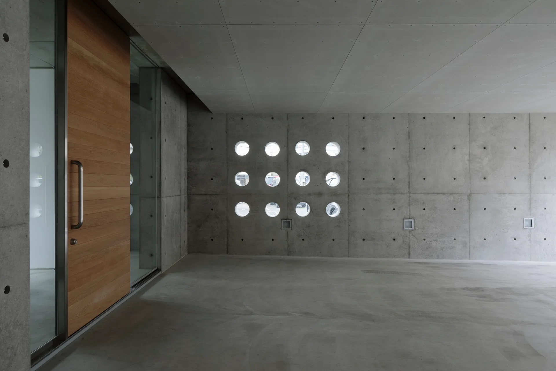 A minimalist concrete entryway features a large wood-paneled door and a grid of twelve circular porthole windows.