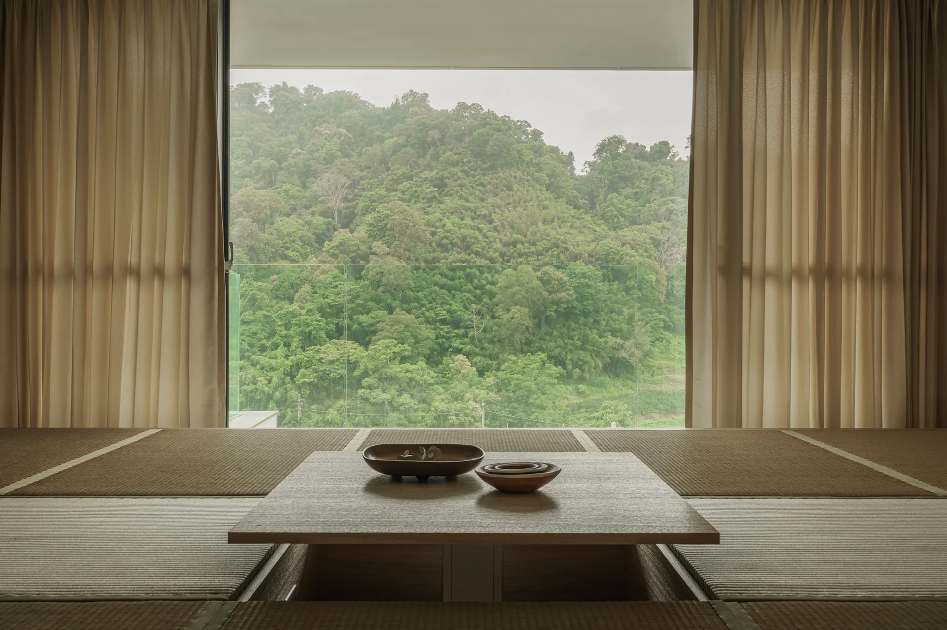 A low wooden table sits on tatami mat flooring before a large picture window showing a lush forest view, framed by beige curtains.