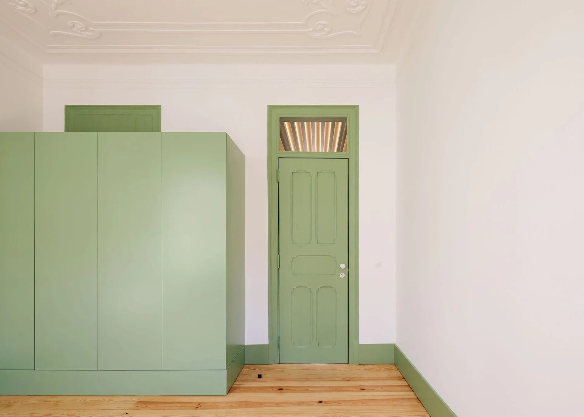 A minimalist room features a tall sage green wardrobe next to a matching green door and baseboard against white walls and light wood floors.