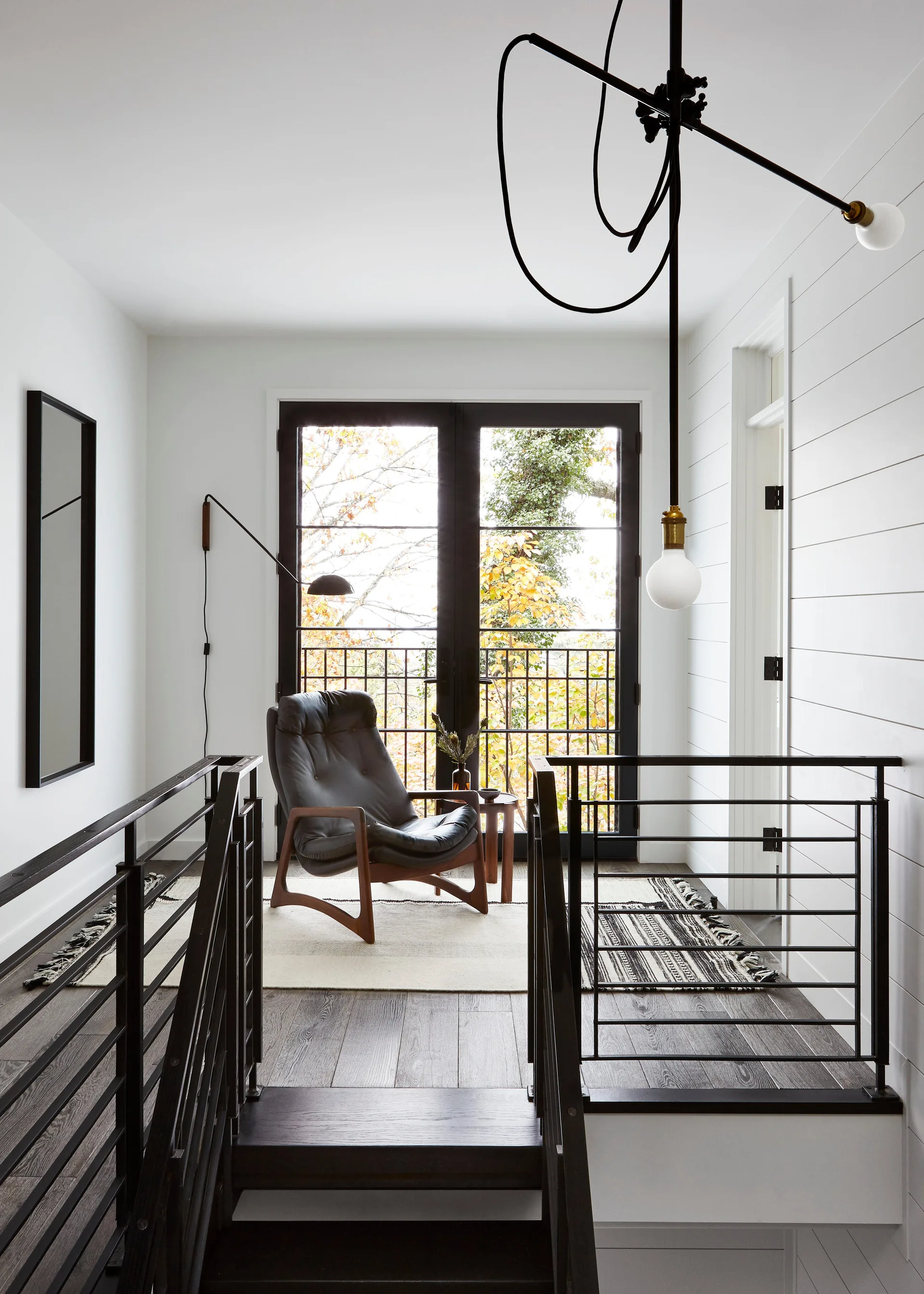 A modern staircase landing featuring a black leather lounge chair on a patterned rug, set before black-framed glass doors and white shiplap walls.