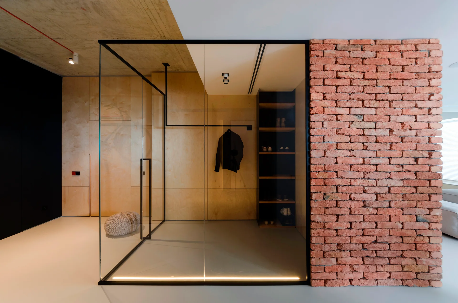 A modern glass-enclosed walk-in closet is flanked by light plywood paneling and an exposed red brick wall.