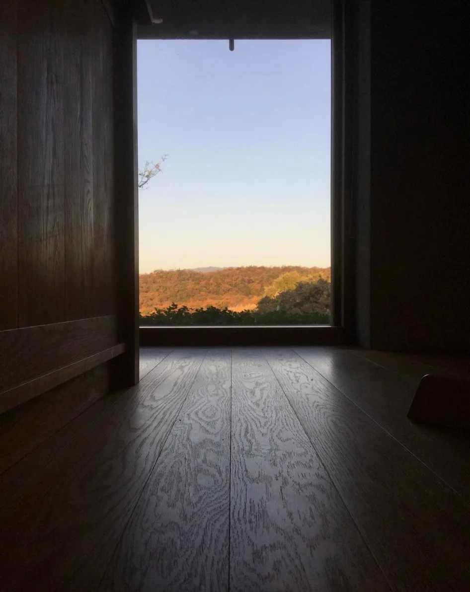 A low-angle view of textured wood floorboards and wood wall paneling leading to a large square window framing a sunny hillside landscape.