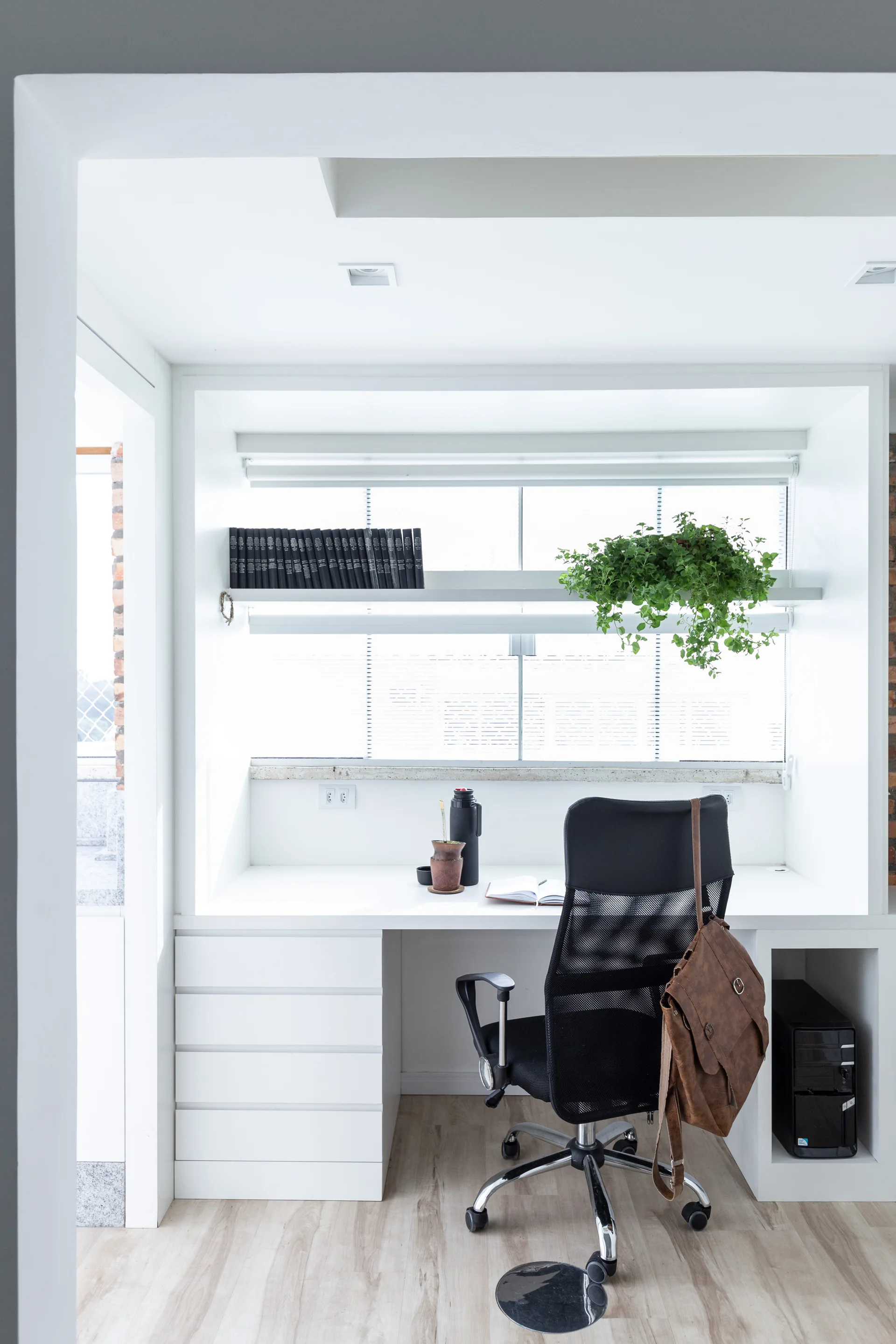 A modern home office with a built-in white desk, a black mesh chair, and a white floating shelf containing books and a trailing plant in front of a window.