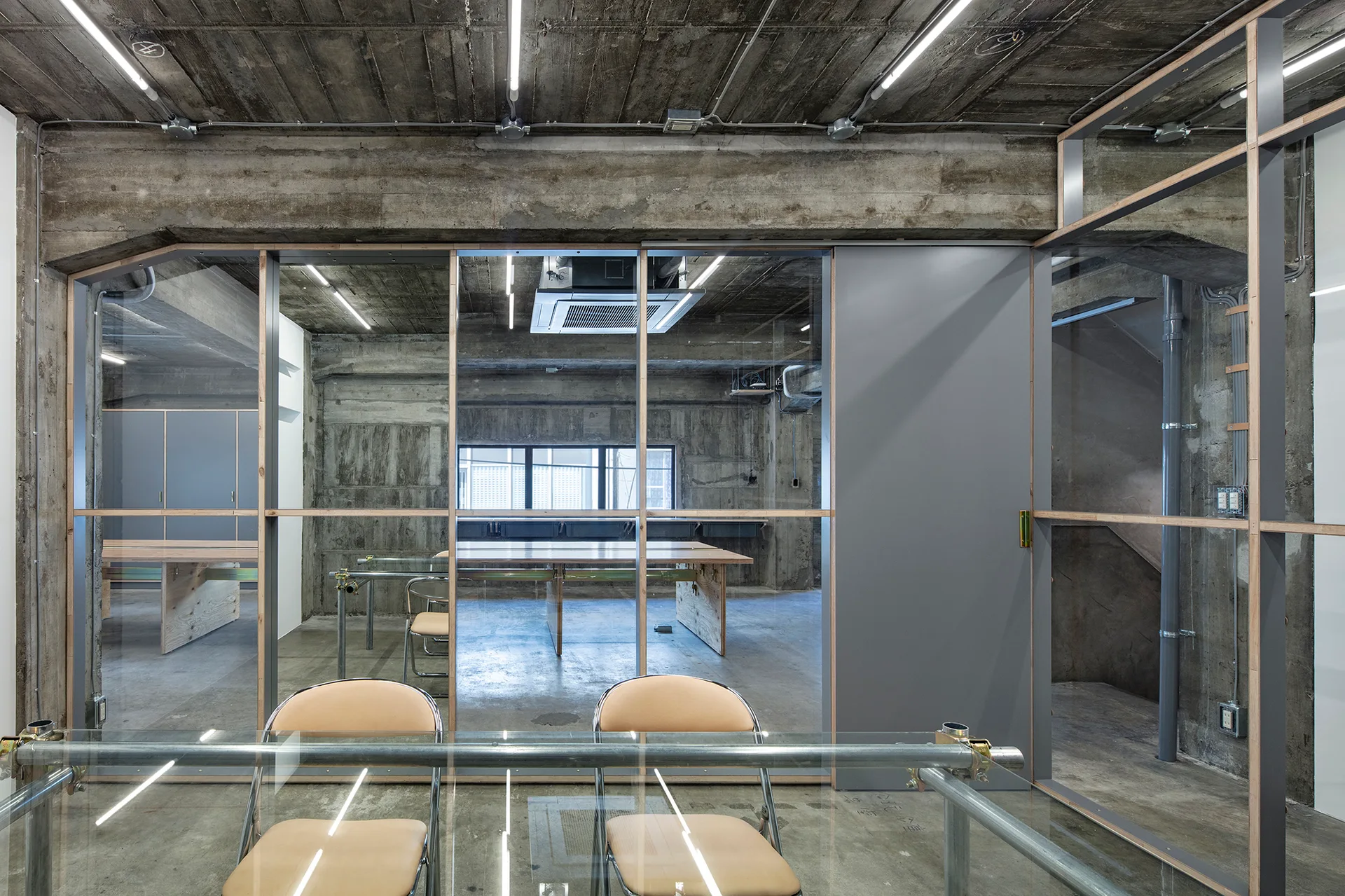 An industrial office space features raw concrete walls, glass partitions with wood framing, and metal-framed tables with tan chairs.