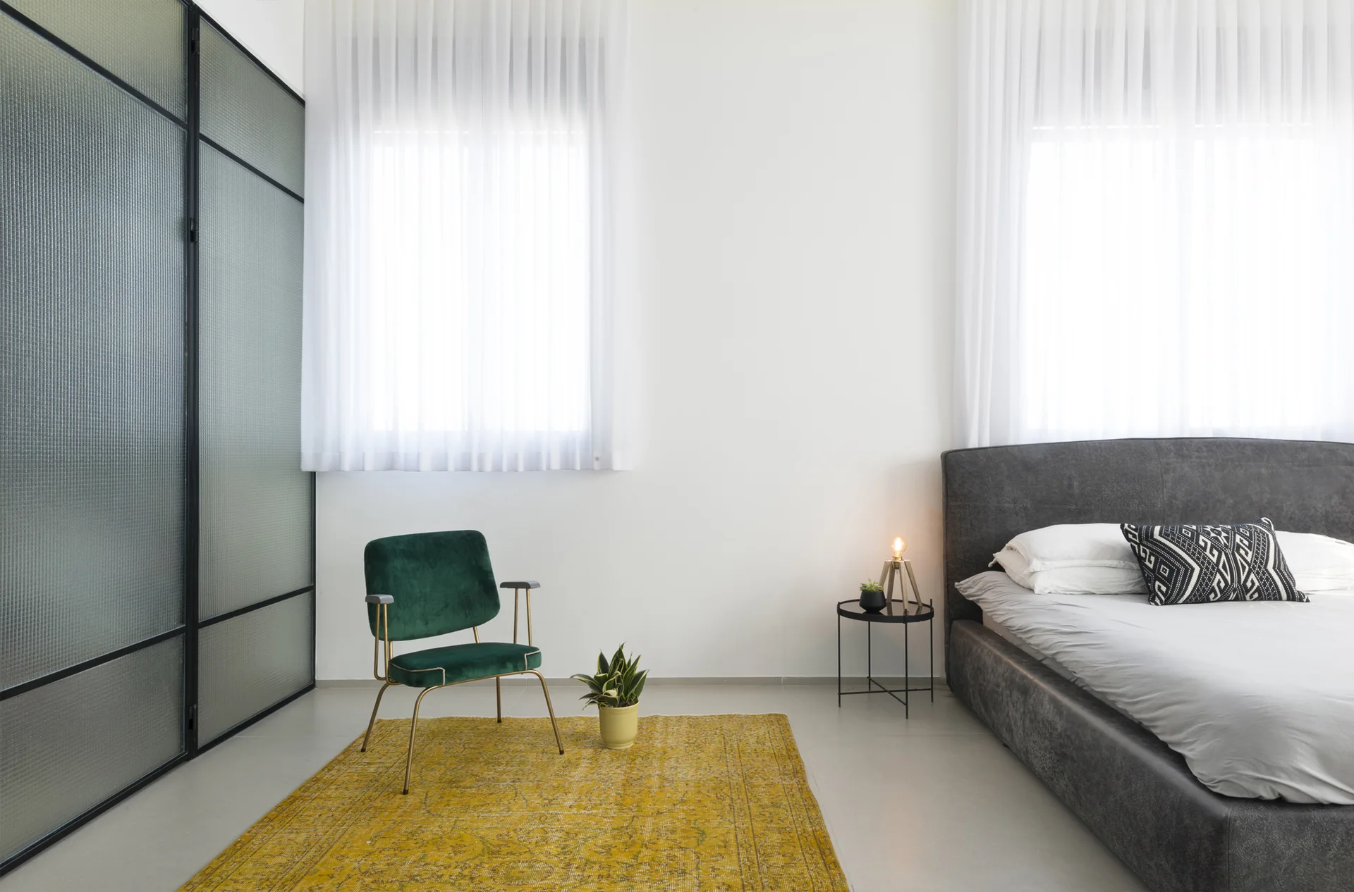 A minimalist bedroom featuring a dark gray upholstered bed next to a green velvet accent chair on a yellow area rug.