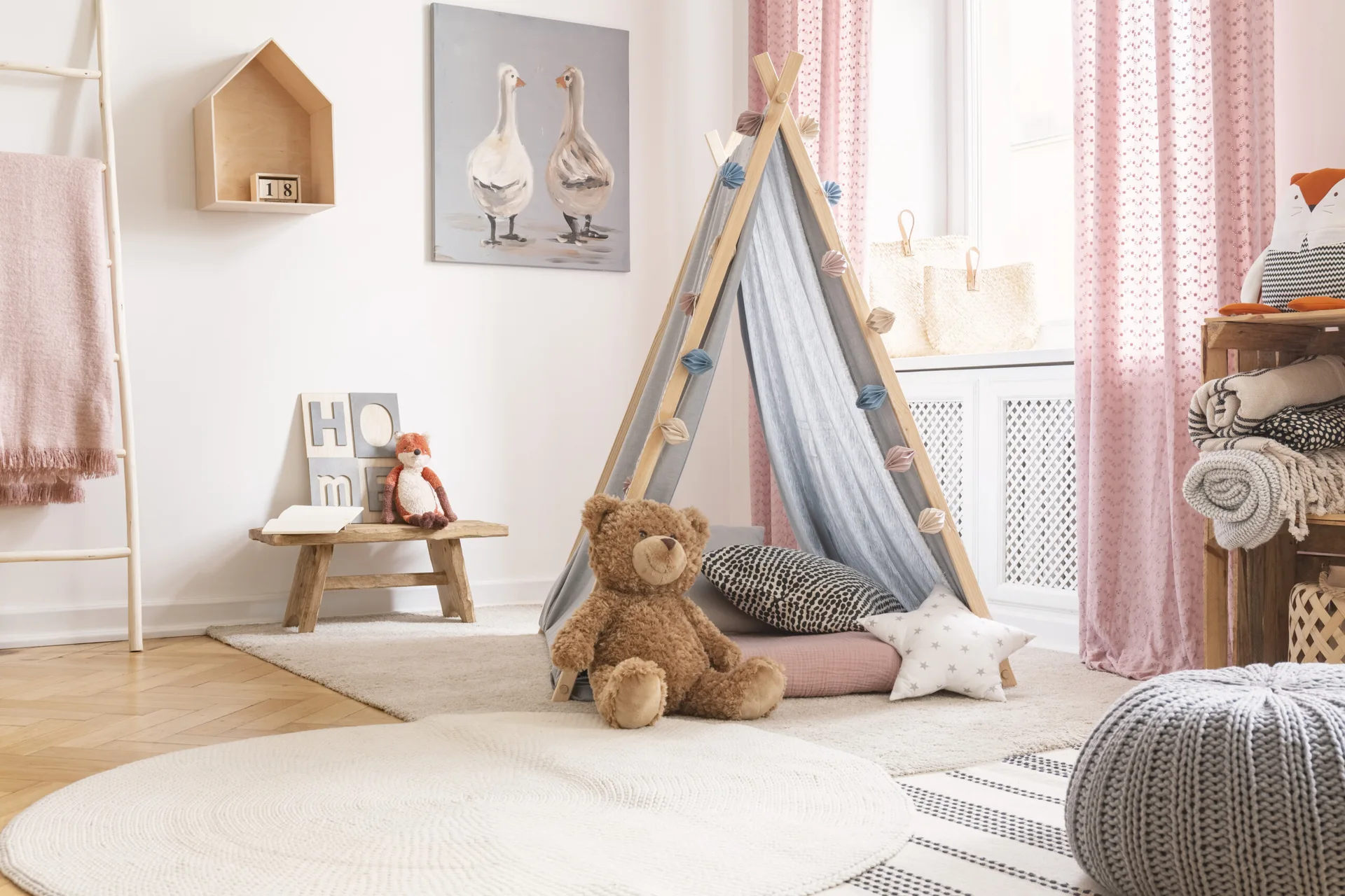 A kids playroom features a fabric teepee tent, a large teddy bear, pink patterned curtains, and layered rugs on a herringbone wood floor.