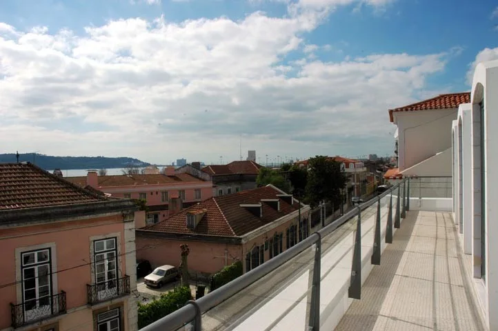 A long, narrow balcony with white tiled flooring and a grey metal and glass railing overlooks a city with terracotta rooftops and a distant waterway.