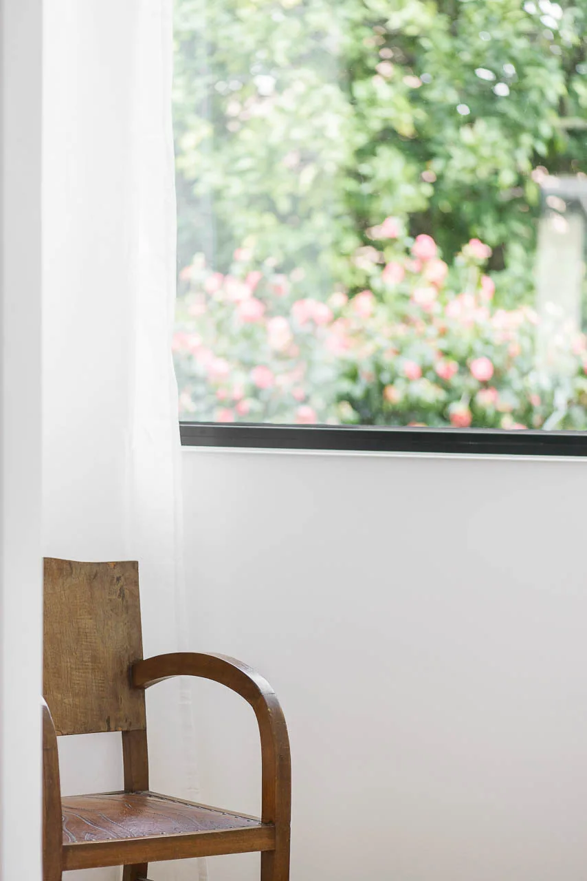 A vintage wooden armchair sits in a white corner next to a window with white curtains showcasing a green garden.