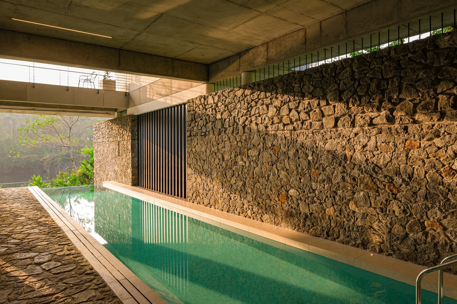 An open-air lap pool with teal water is flanked by a textured natural stone wall under an exposed concrete ceiling.