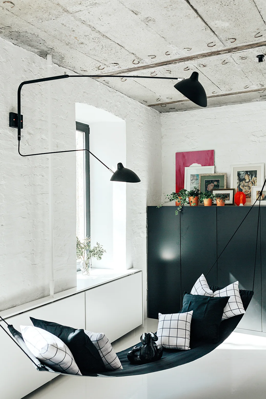 A black fabric hammock with grid-patterned pillows hangs in a bright room with white brick walls and an exposed concrete ceiling.