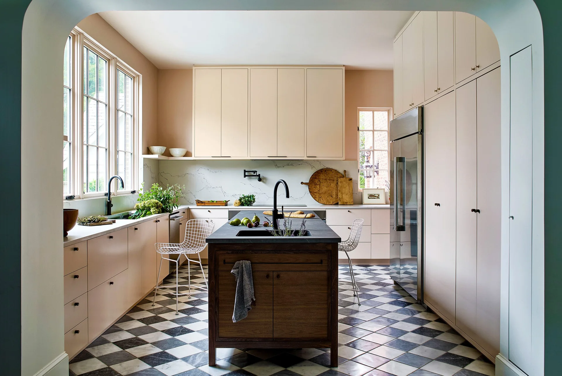 A contemporary kitchen features light pink cabinets, a dark wood central island with a black faucet, and a black-and-white checkerboard tile floor.