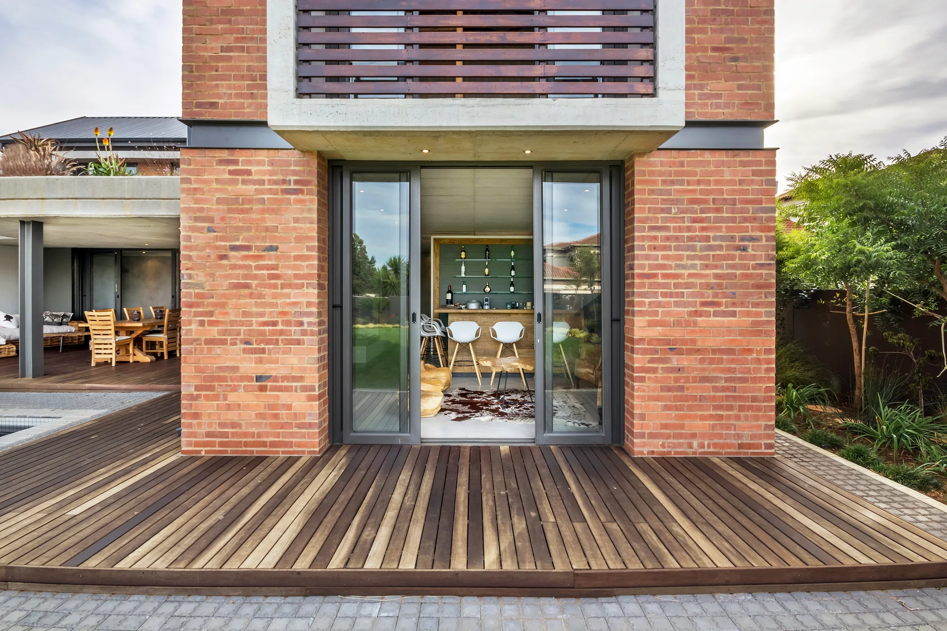 A modern red brick home exterior featuring a wooden deck and sliding glass doors that look into a bar area.