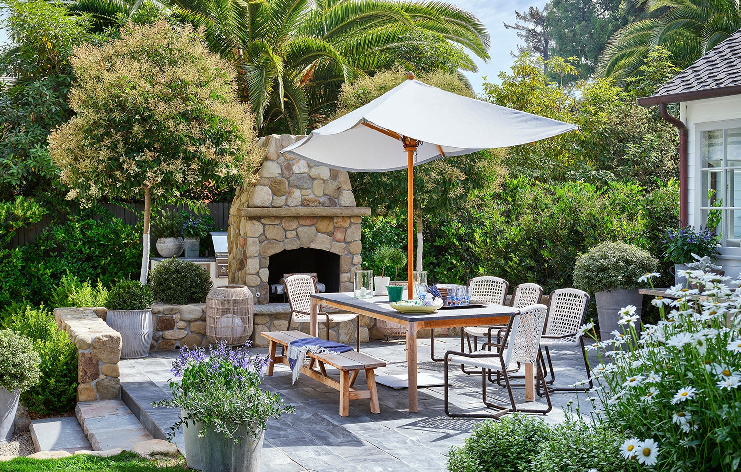 An outdoor stone patio features a large rectangular dining table under a white umbrella, with a rustic stone fireplace in the background.
