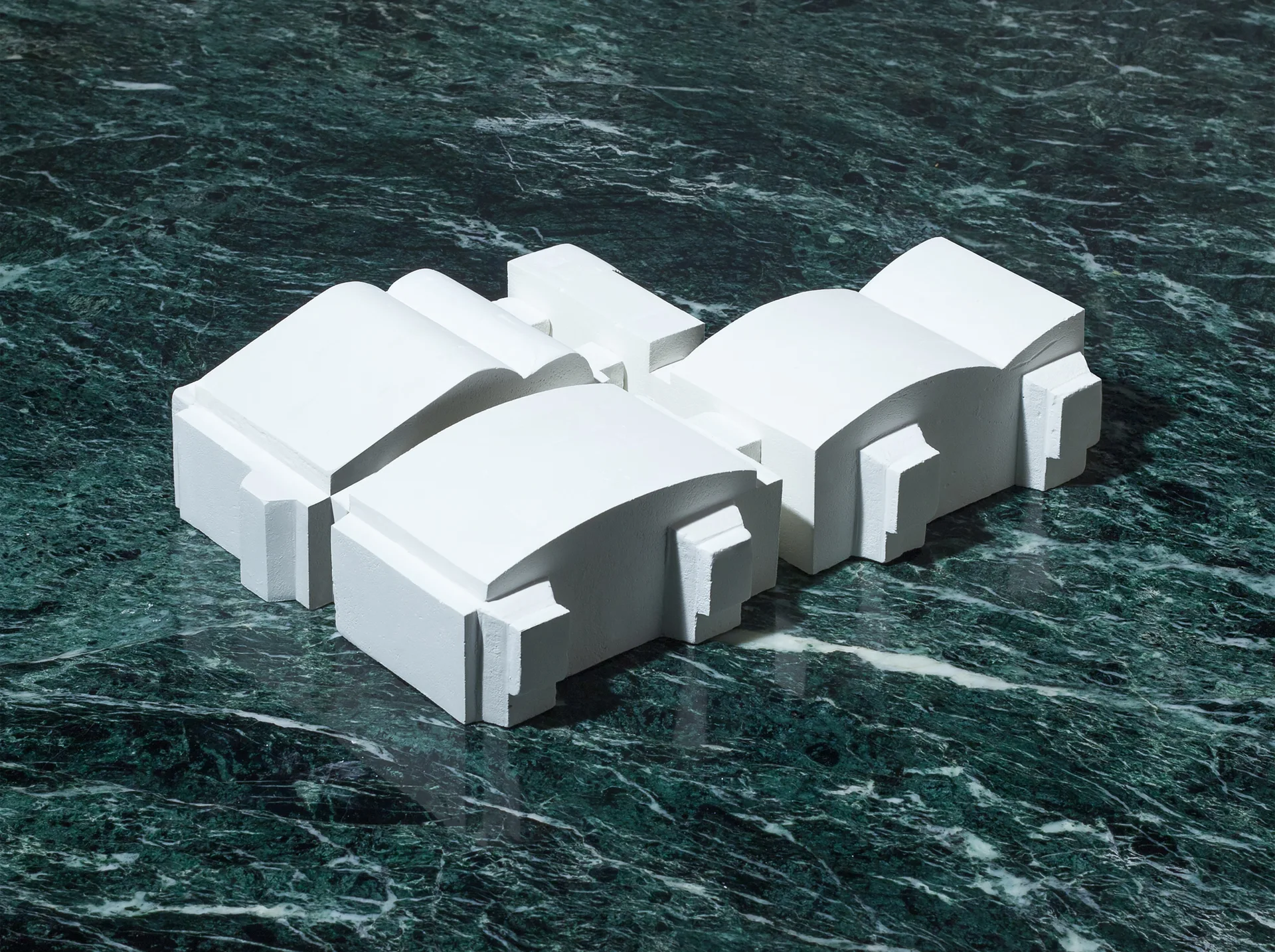 Three matte white geometric architectural components rest on a polished surface of dark green veined marble.