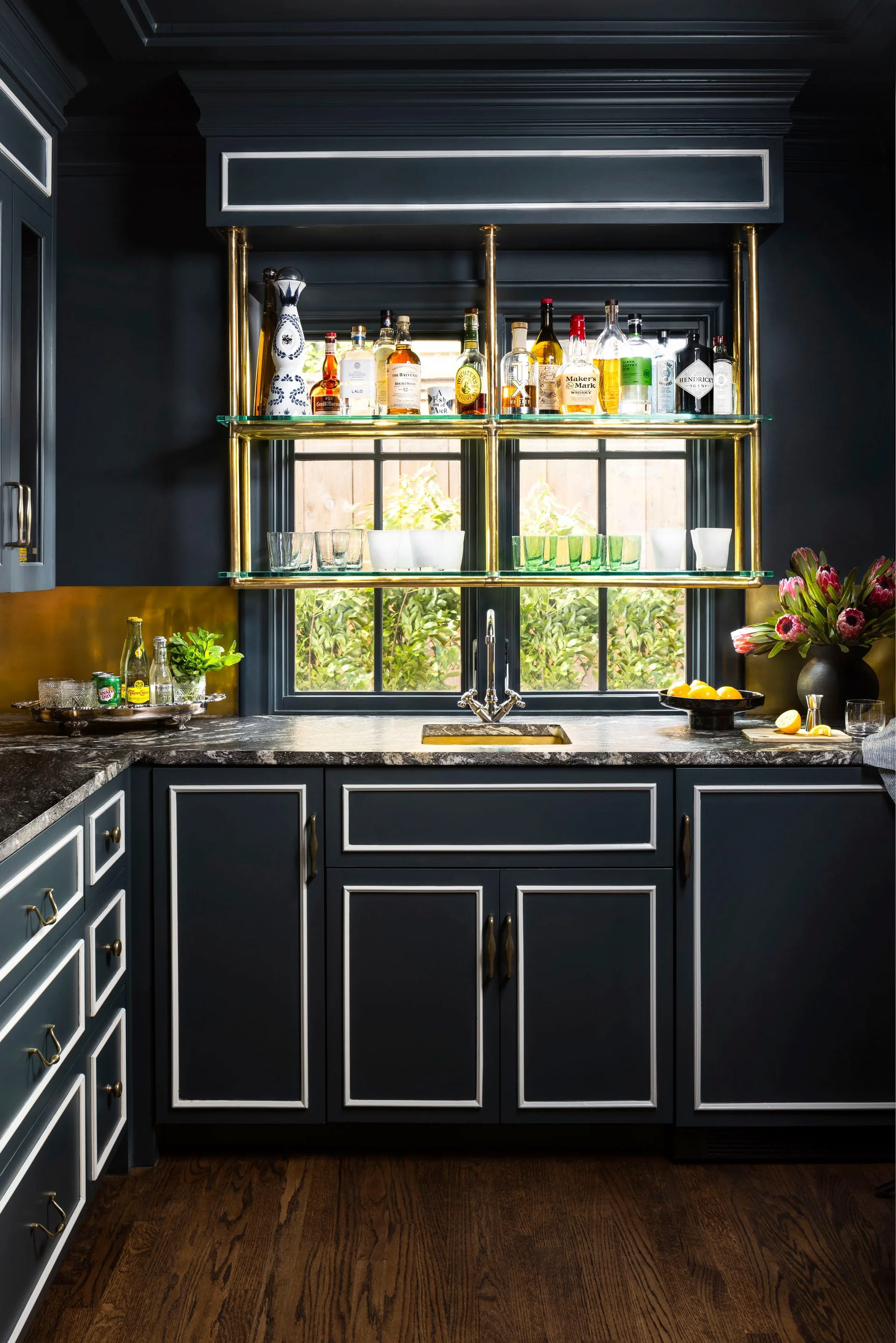 A moody navy wet bar featuring white-trimmed cabinetry, dark marble countertops, and a brass-framed glass shelf suspended over a window.
