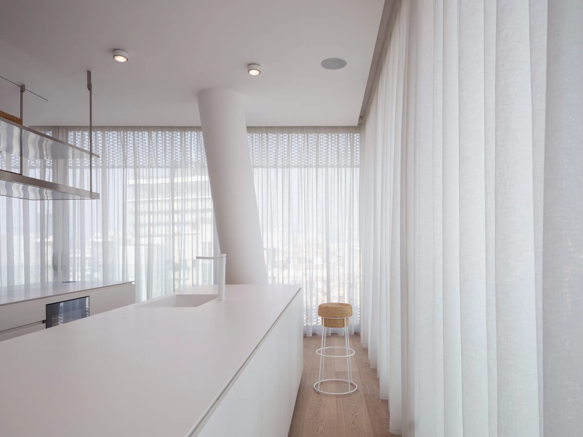 A minimalist white kitchen island with an integrated sink sits on wood flooring, surrounded by floor-to-ceiling sheer white curtains.