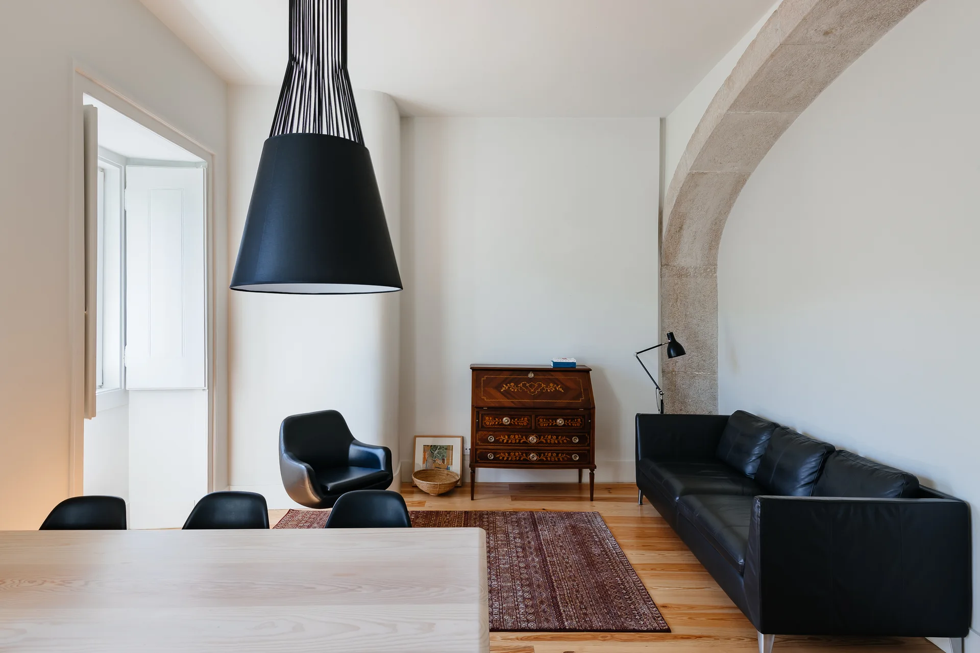 A minimalist living room features a black leather sofa and a vintage secretary desk set beneath a prominent architectural stone archway.