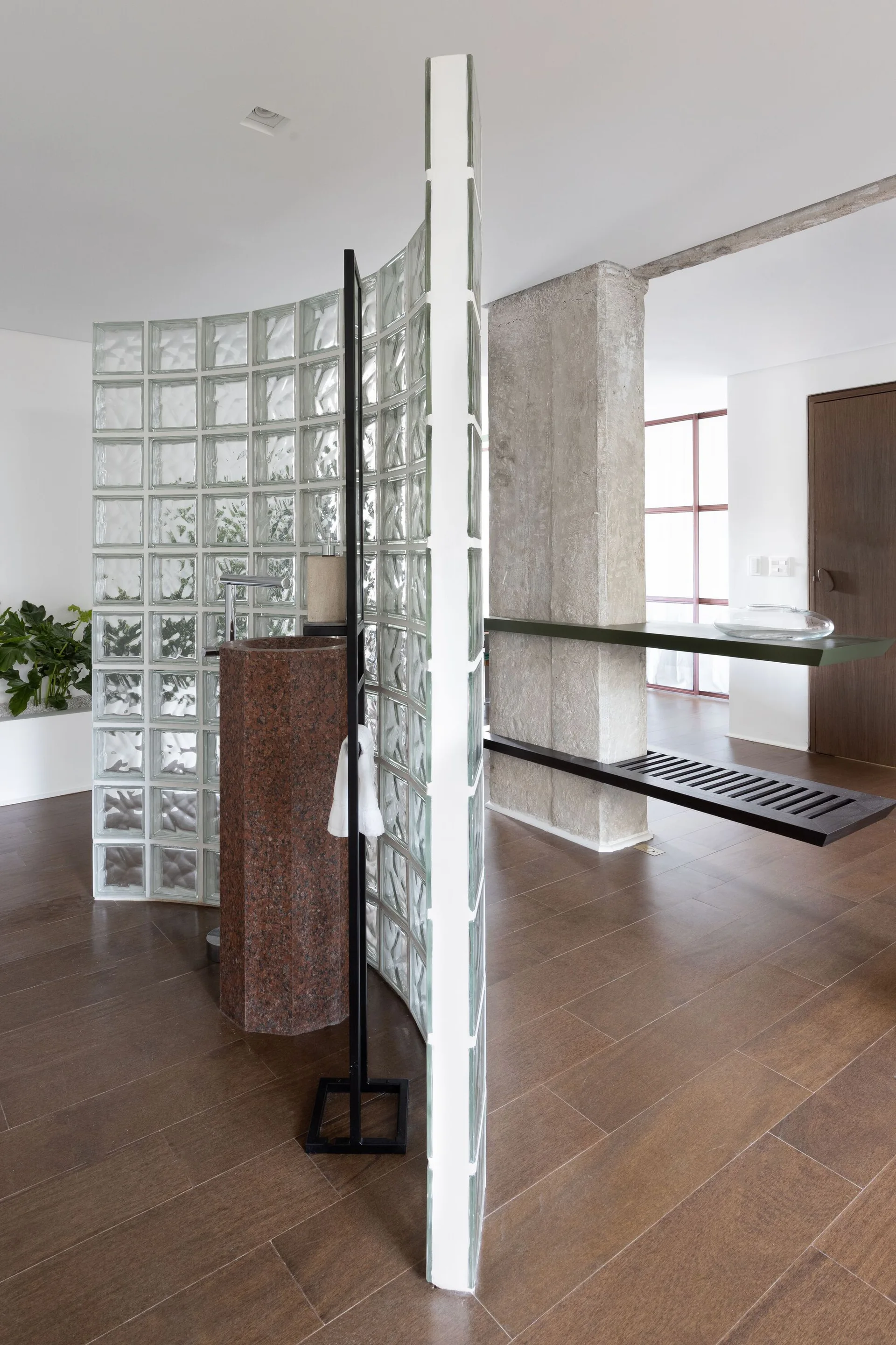 A modern bathroom area featuring a curved glass block partition wall next to a dark stone pedestal sink and a raw concrete column.
