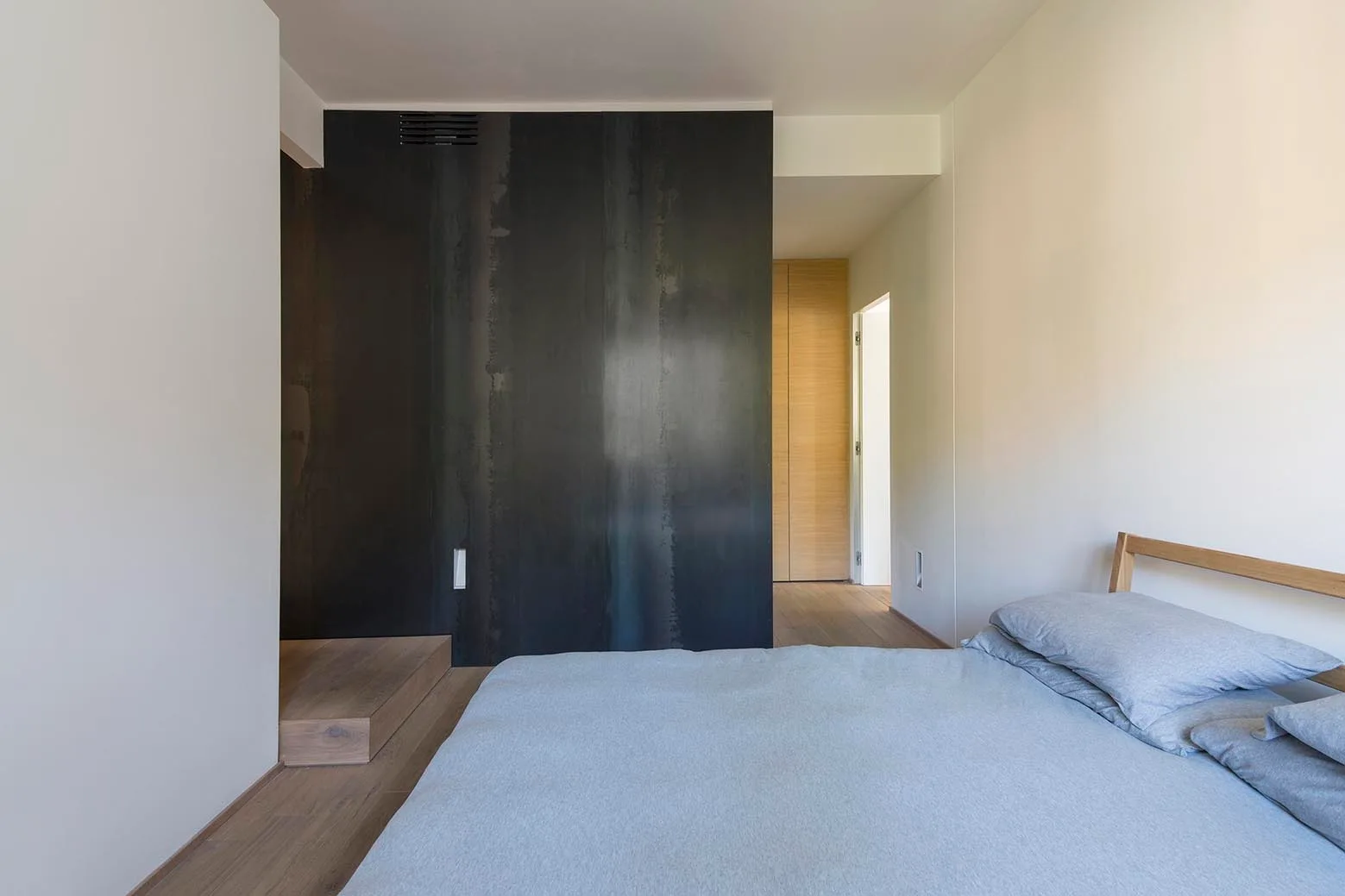 A minimalist bedroom featuring a wooden bed with gray bedding, light wood floors, and a dark metal partition wall.