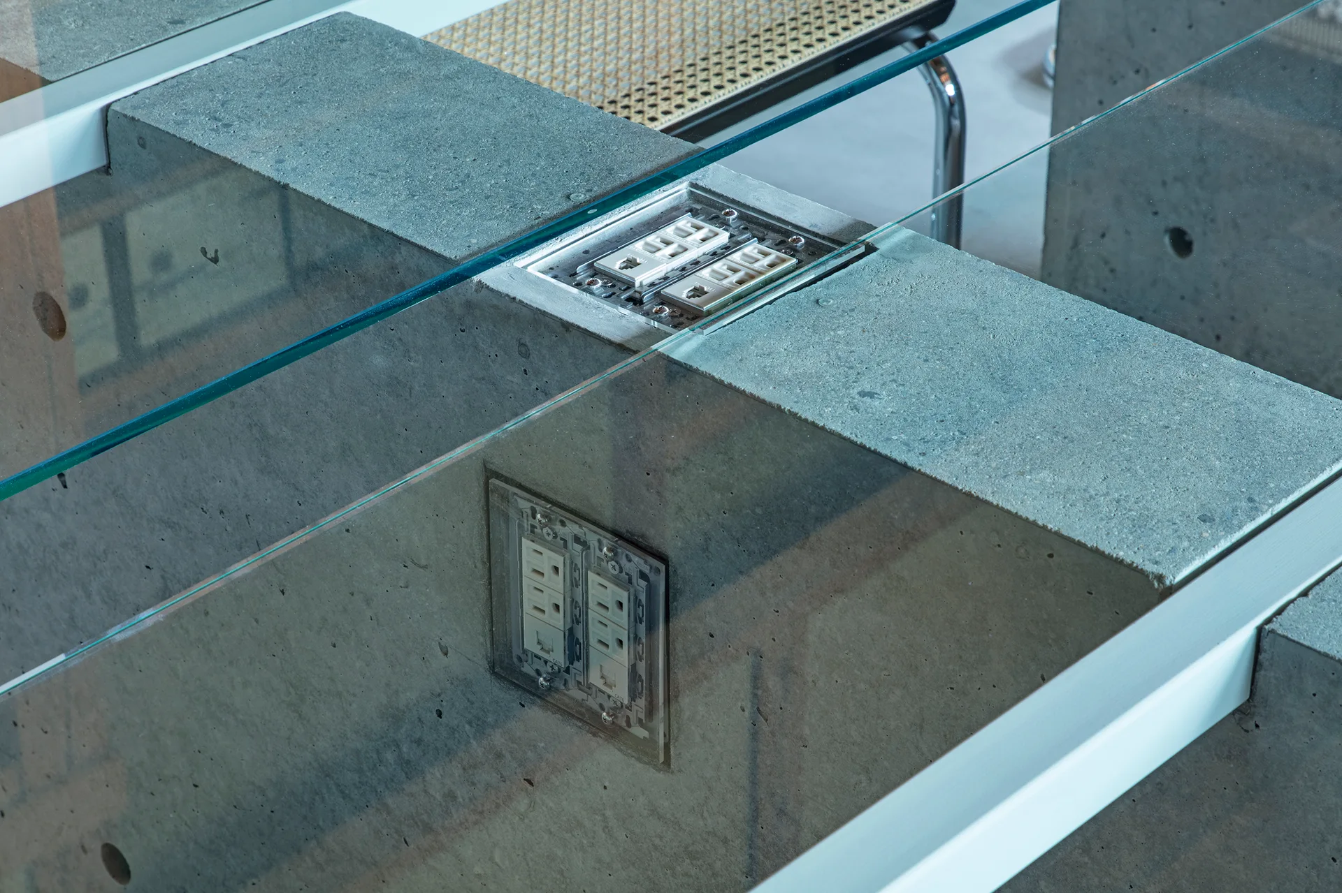 Close-up of a custom raw concrete table featuring integrated electrical outlets in the base beneath a clear glass top.