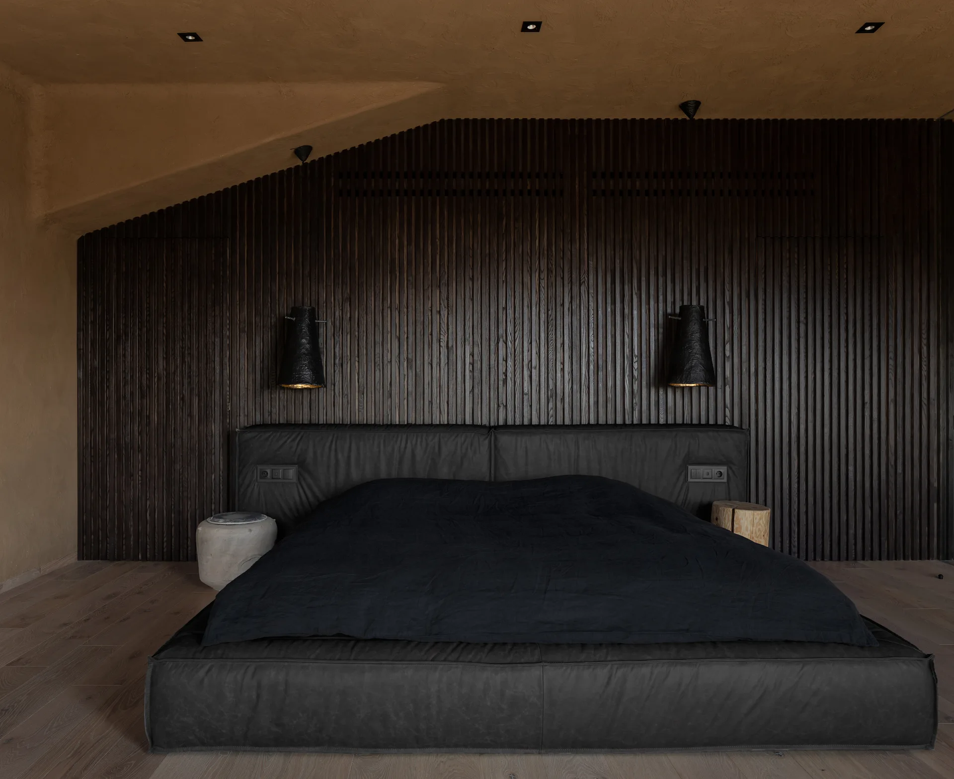 A low-profile black bed is set against a dark wood slatted wall under a sloped tan ceiling, flanked by small pedestal nightstands and dark wall lamps.