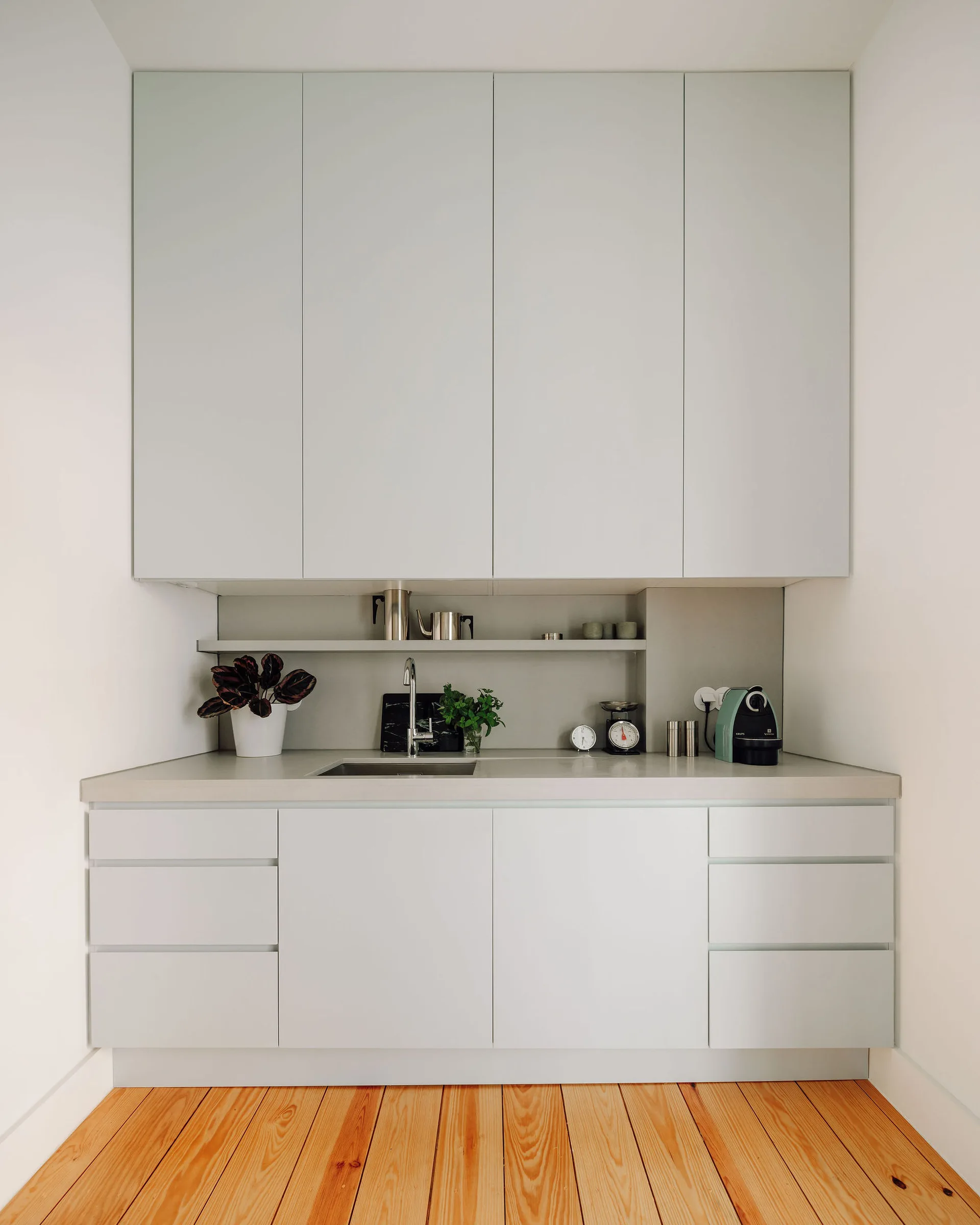 A minimalist light grey kitchenette featuring handleless cabinets, a small undermount sink, and natural wood plank flooring.