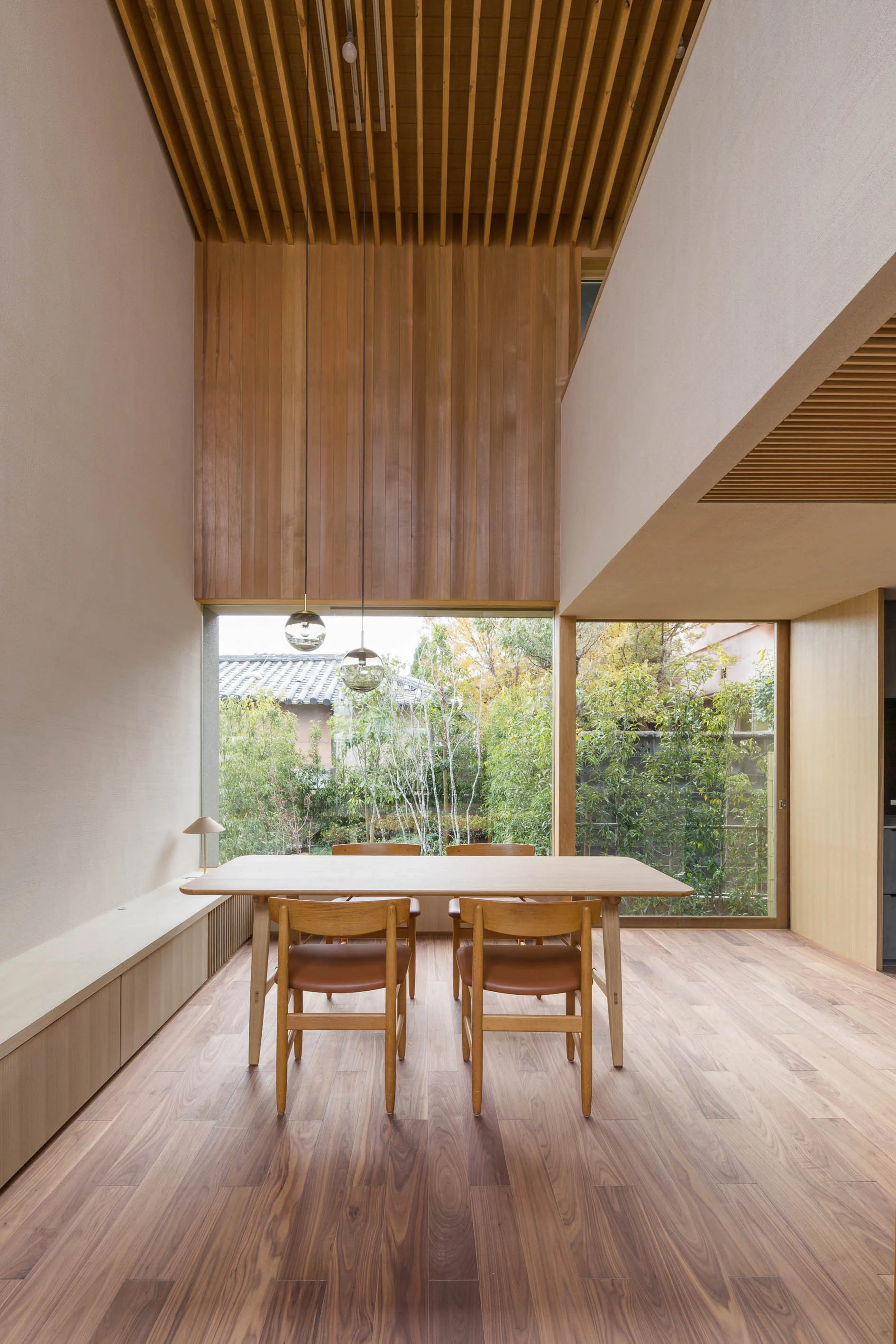 A light wood dining table and chairs sit in a double-height room with a slatted wood ceiling and large windows looking out to a garden.