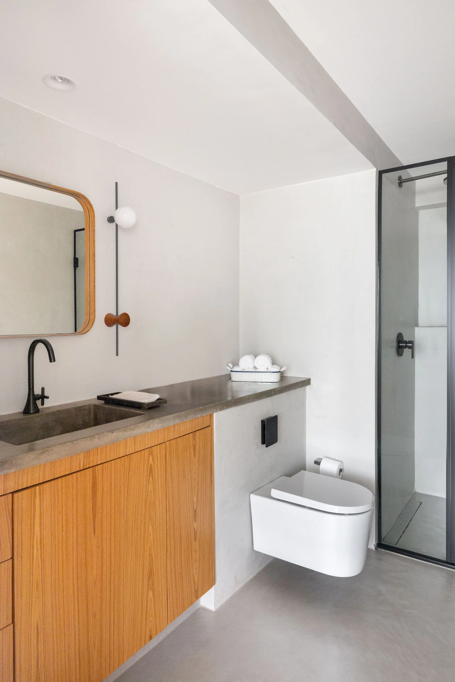 A modern bathroom with a wood cabinet vanity, integrated concrete sink, black faucet, wall-mounted toilet, and glass shower enclosure.