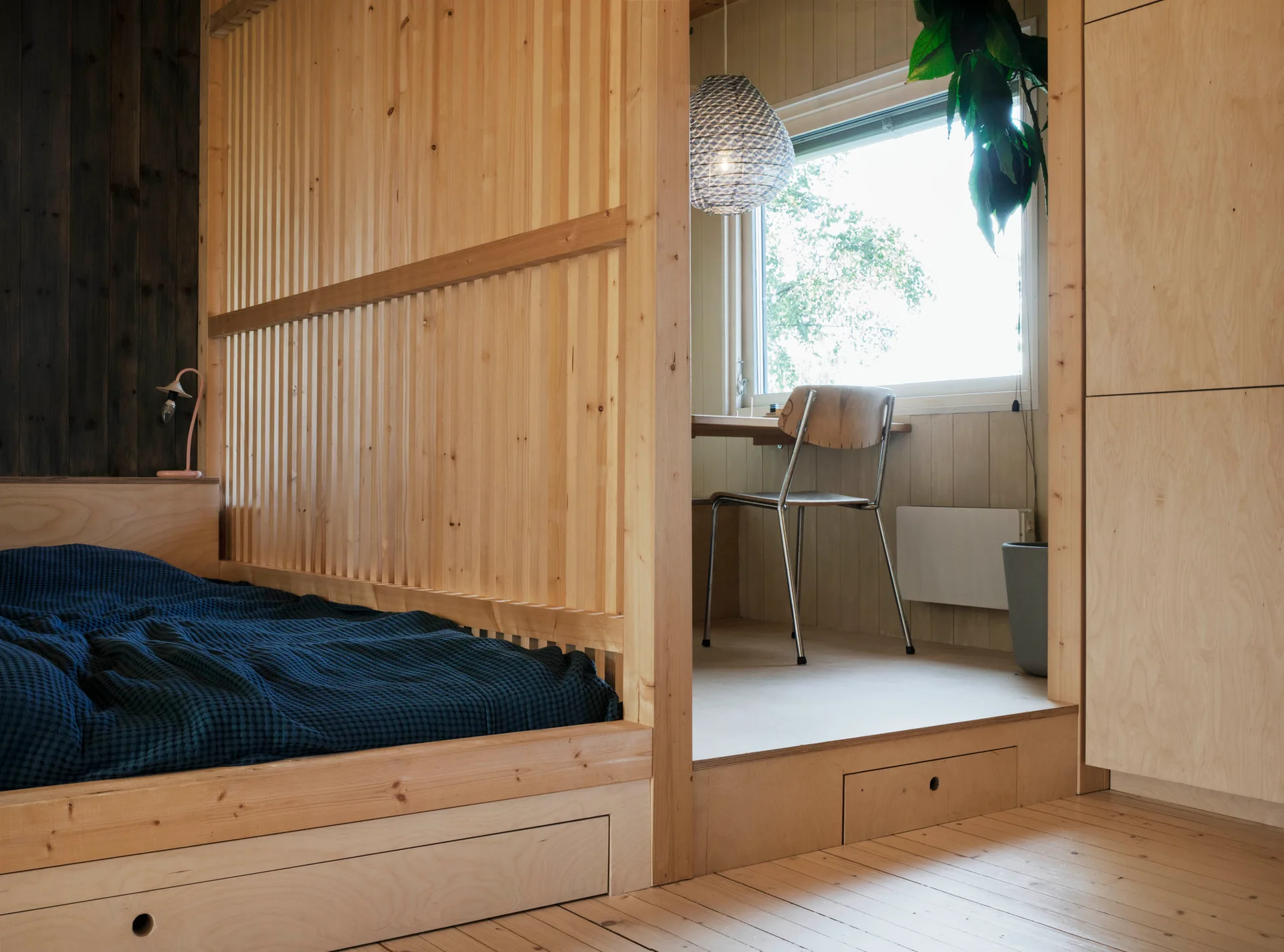 A platform bed with a dark blue cover is separated from a raised desk nook by a light wood slat partition.