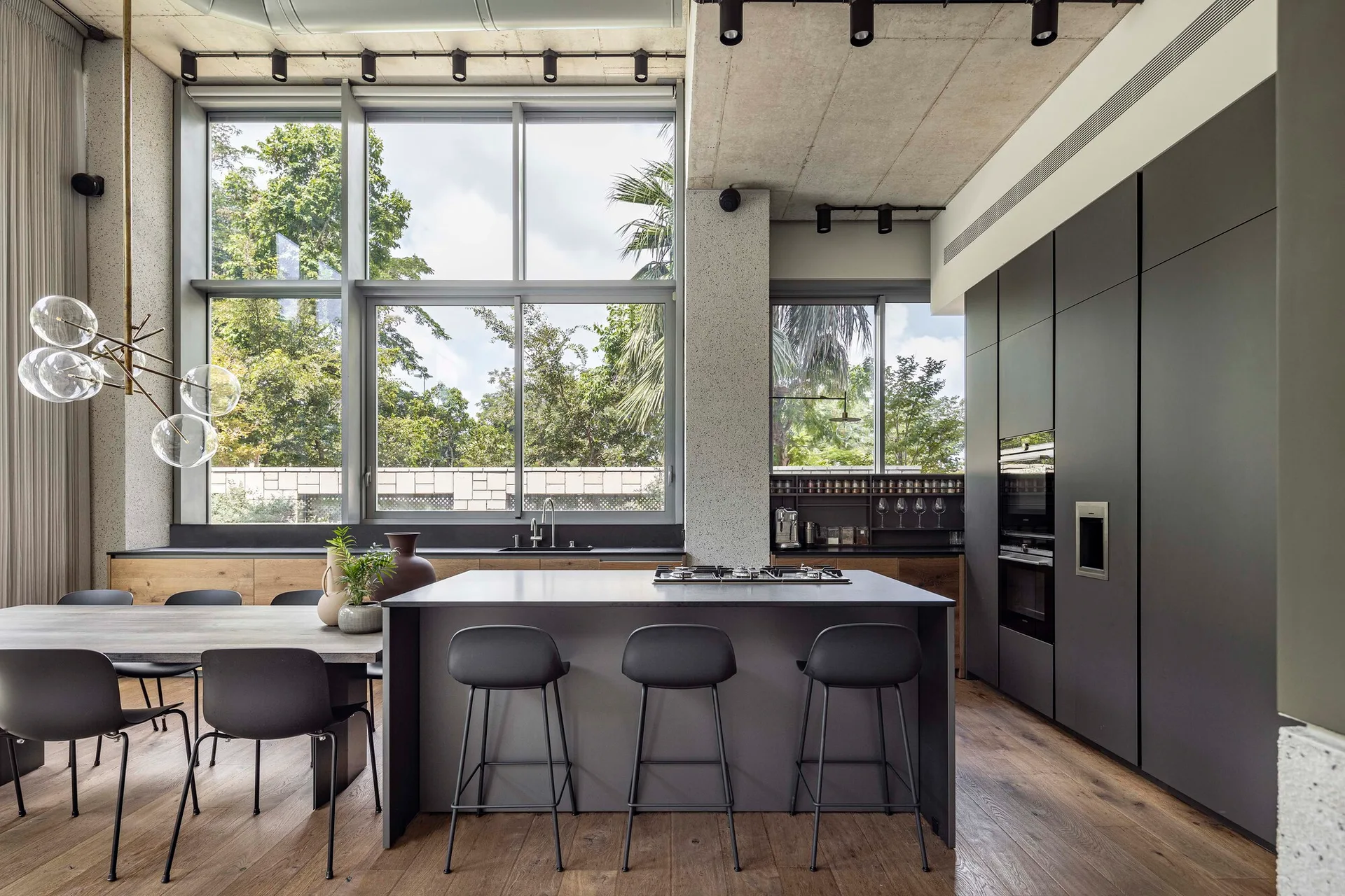 A modern kitchen and dining space features a charcoal island with stools, tall dark cabinets, an exposed concrete ceiling, and large windows.