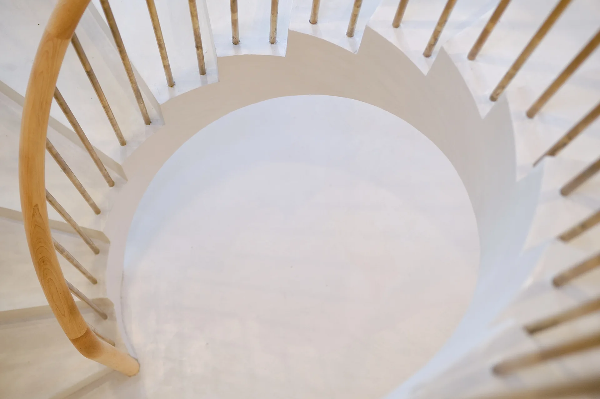 A top-down view of a modern white spiral staircase featuring a curved light wood handrail and matching slender balusters.