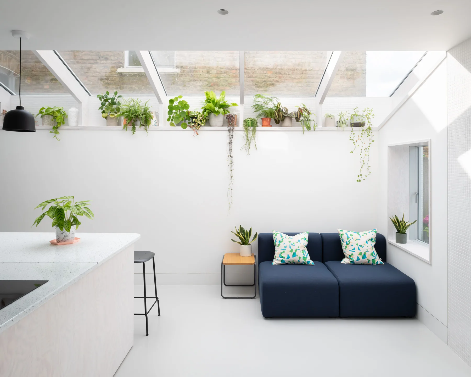 A bright sunroom features a navy blue modular sofa beneath a long plant shelf filled with potted greenery under a sloped skylight.