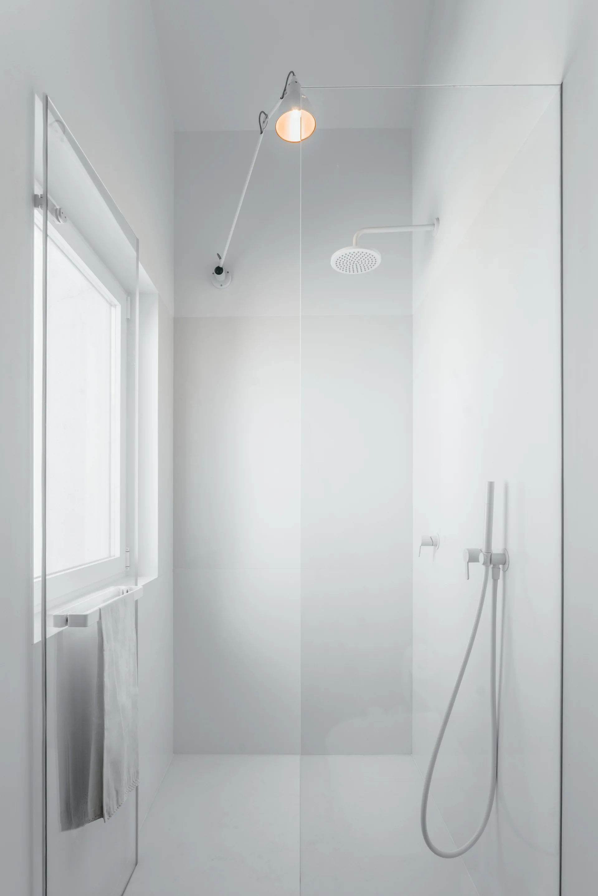 A minimalist white tiled walk-in shower features matching white shower fixtures, a glass panel partition, and a single ceiling-mounted warm spotlight.
