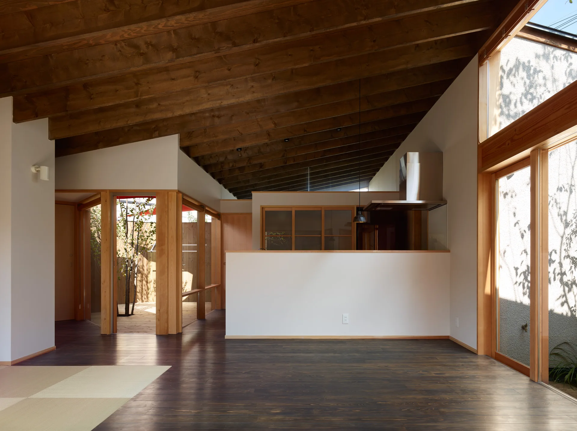 A minimalist open-plan living room features dark wood floors, exposed ceiling beams, and a white partition wall leading to a kitchen.