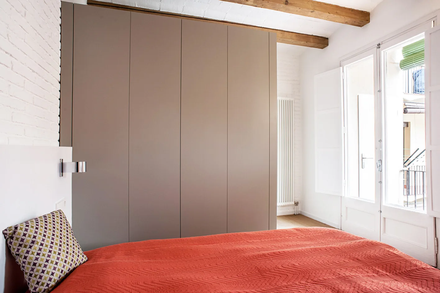 A contemporary bedroom featuring a large matte taupe built-in wardrobe, a bed with coral bedding, and exposed wood ceiling beams.