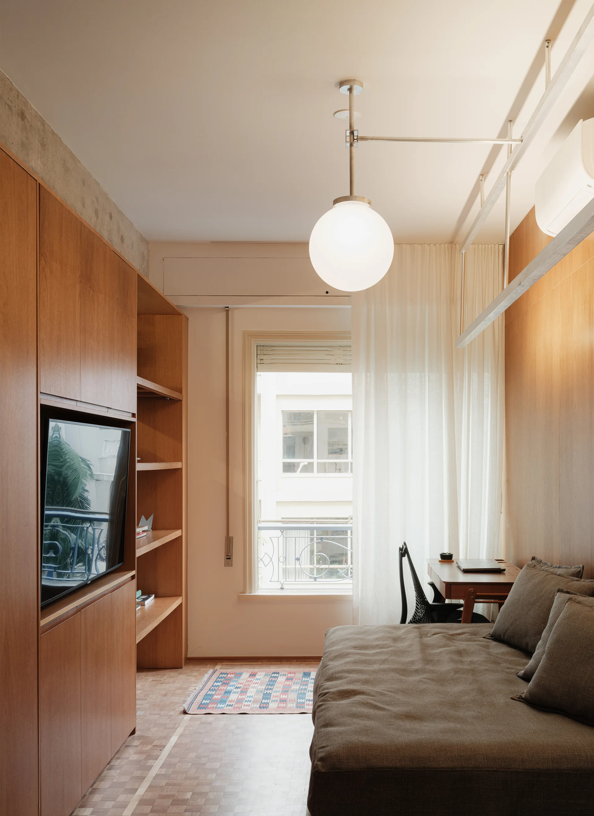 A compact bedroom features custom wood cabinetry with a built-in television, a beige bed, parquet floors, and a desk by a window with sheer curtains.
