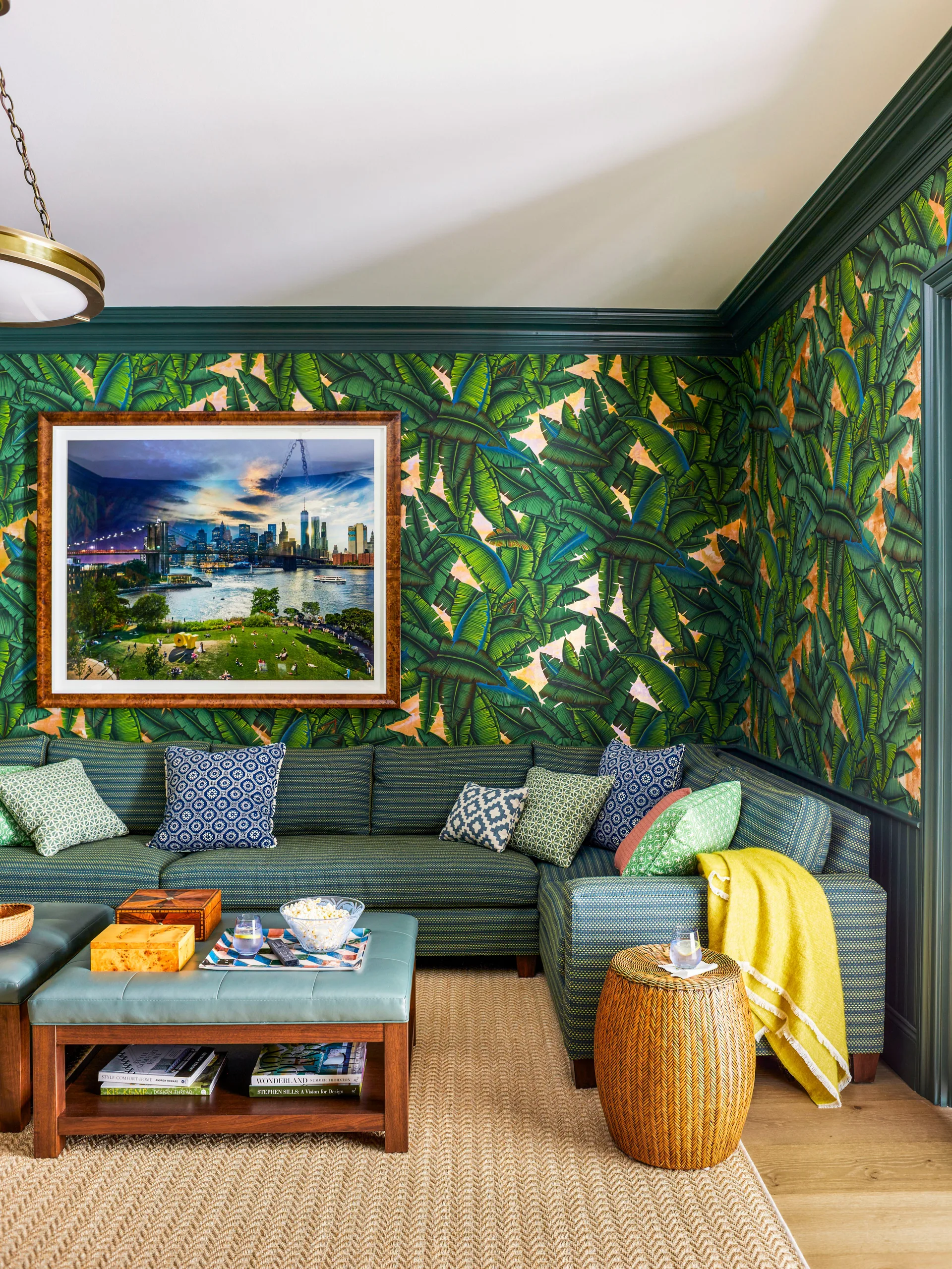 A living room features a patterned teal sectional sofa against bold banana leaf wallpaper with dark green trim and a herringbone woven rug.