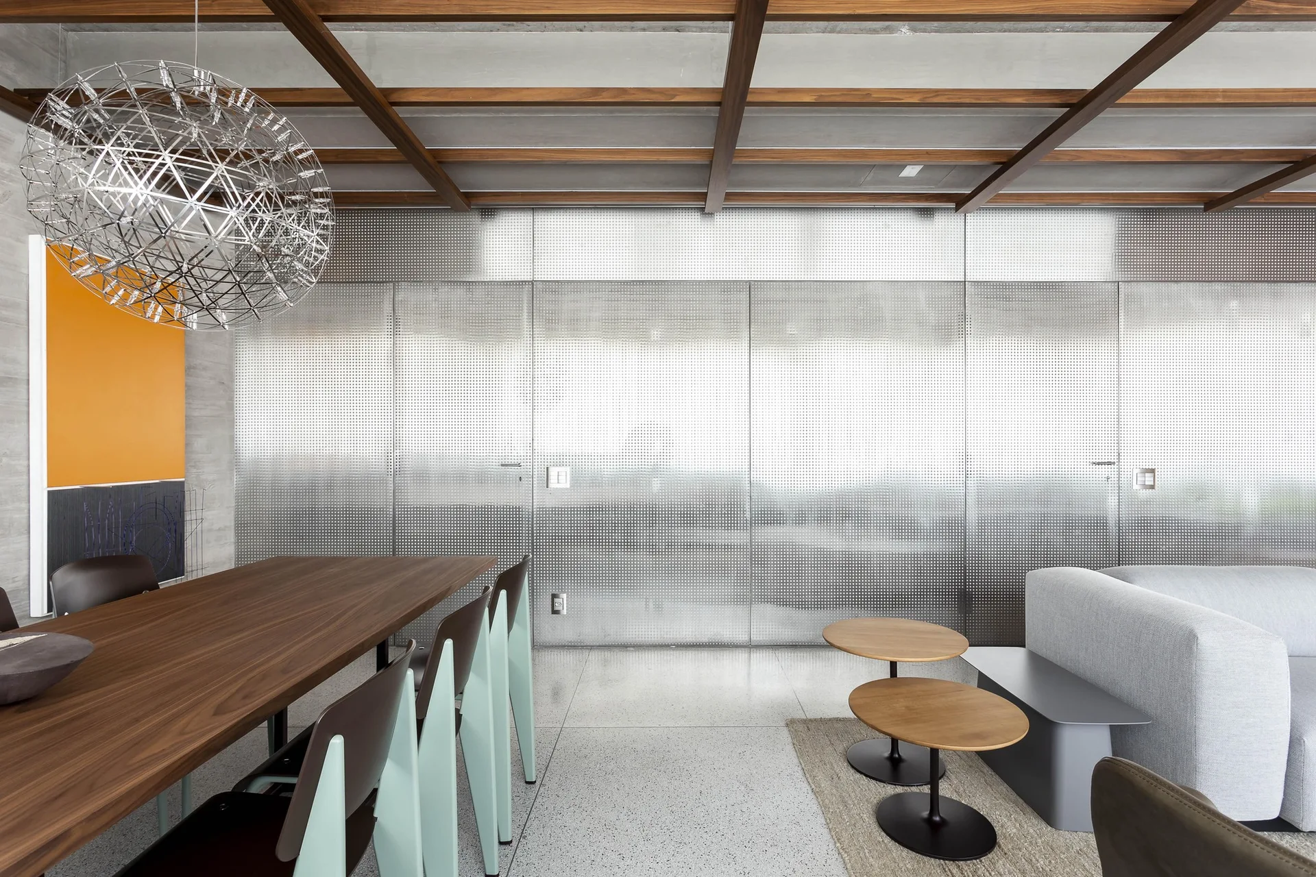 A modern living and dining space featuring perforated metal wall paneling, a wood ceiling grid, and a spherical metal wire pendant light.