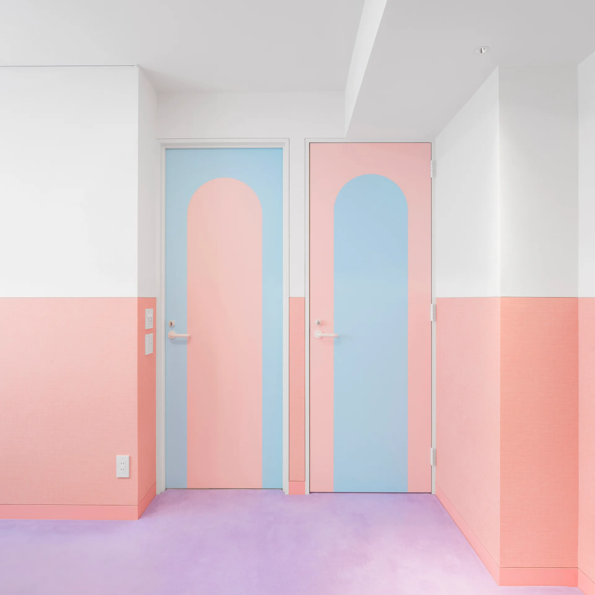 Two interior doors with contrasting pastel pink and blue arched paint designs set against a pink and white two-tone wall with lavender carpet.